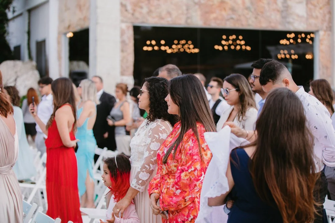 People attending an outdoor event, standing in a row, in front of a building with string lights.