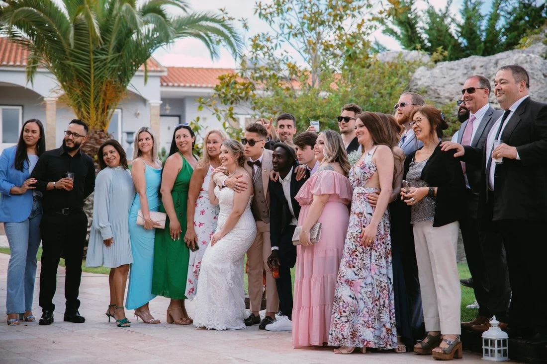 Group of people in formal attire gathered outdoors for a photo, with some holding drinks, under palm trees.