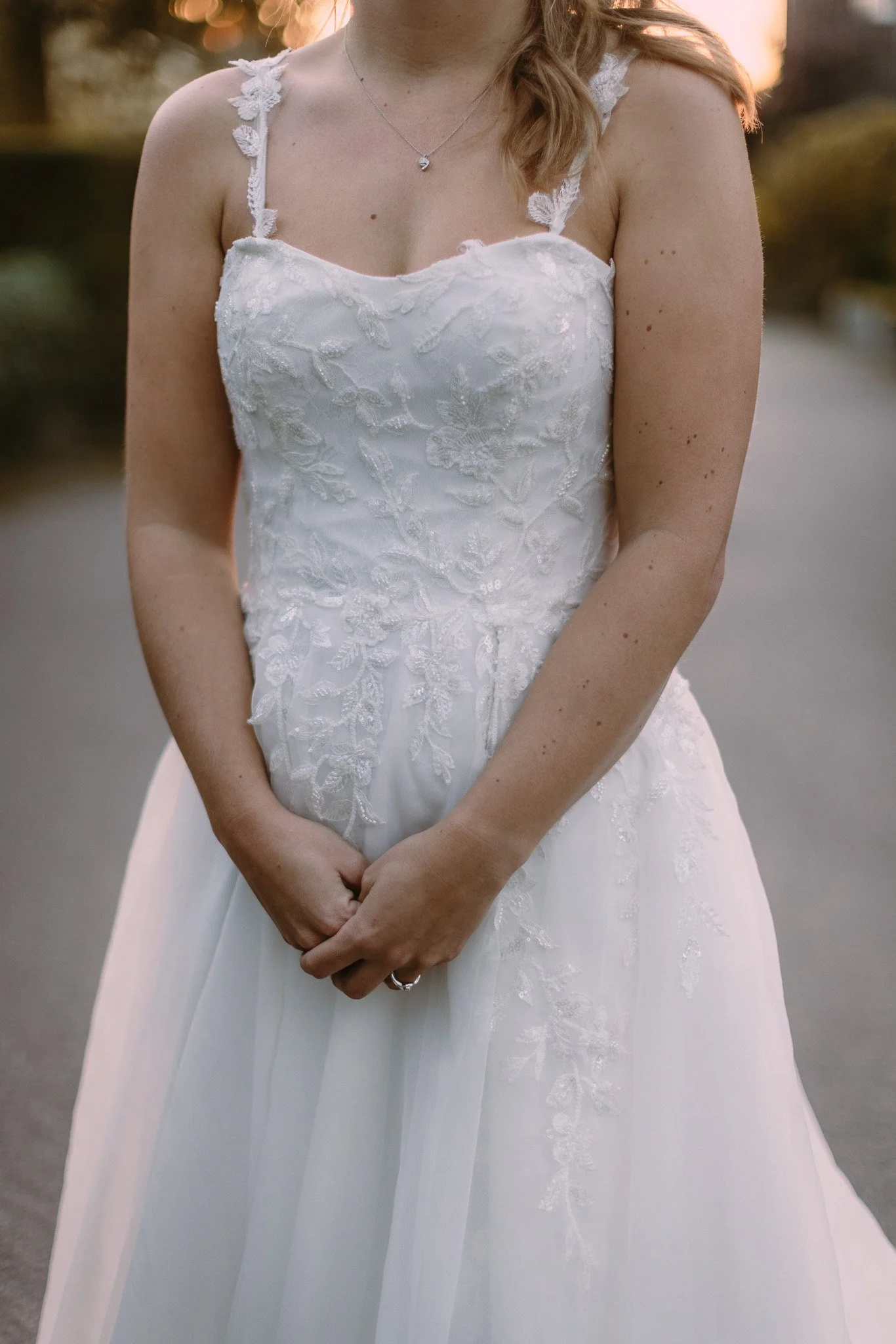A woman in a white wedding dress with lace details, standing outdoors during sunset, with her hands clasped together in front of her.
