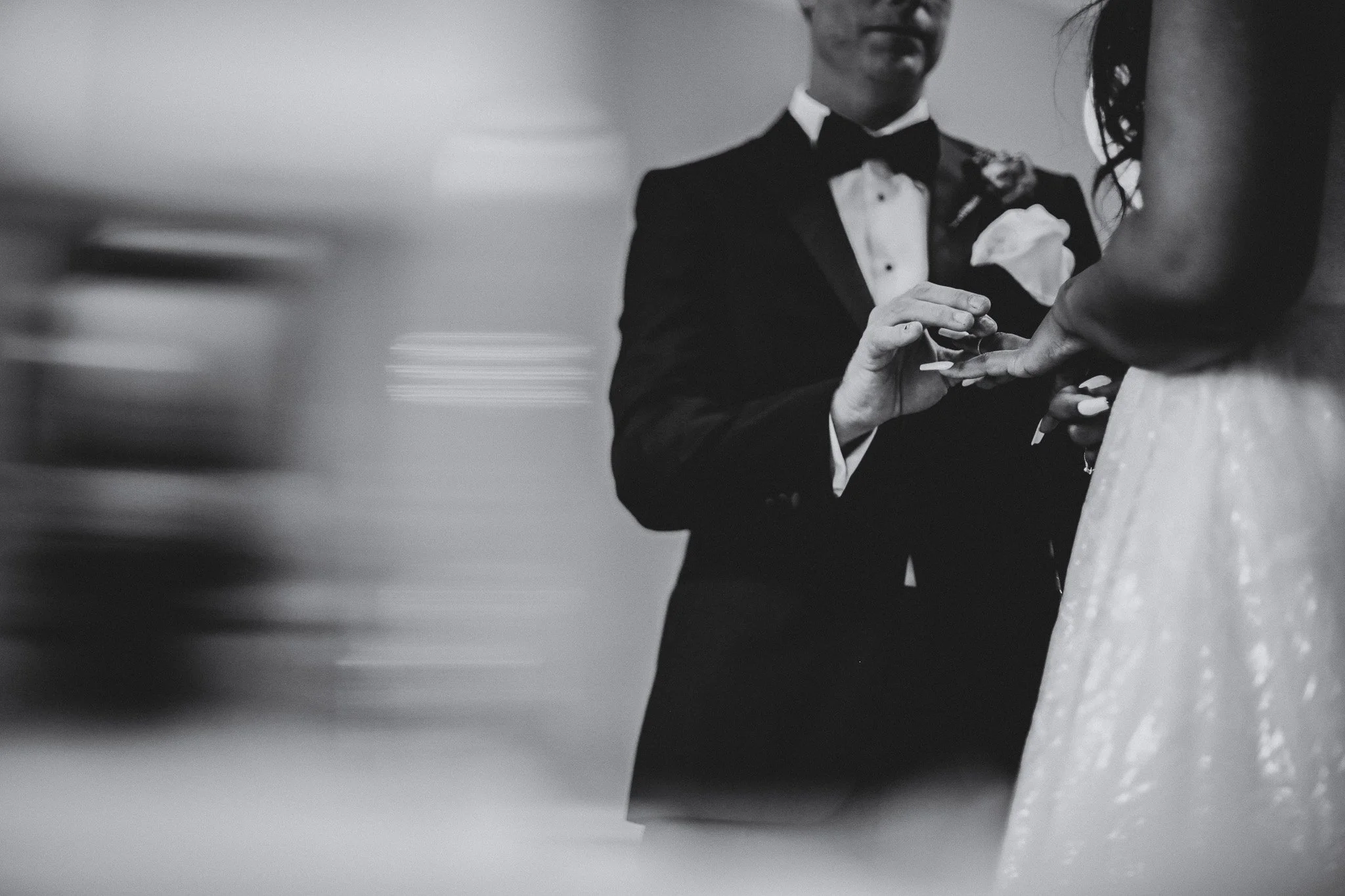 A bride and groom holding hands during their wedding ceremony, with the groom in a tuxedo and the bride in a wedding dress, in black and white.