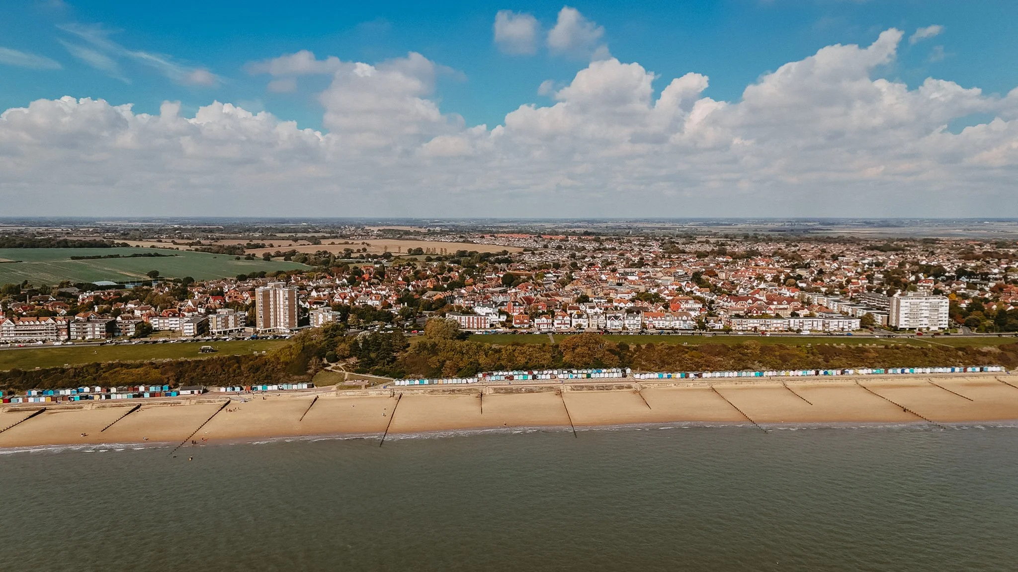 Aerial view of a coastal city with houses, apartment buildings, and green fields under a partly cloudy sky, with a sandy beach and a sea wall in the foreground.