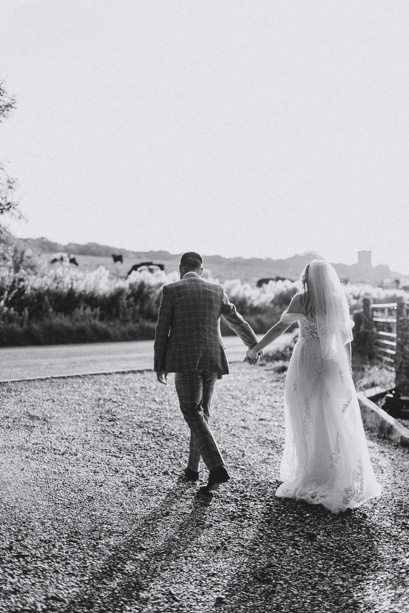 A bride and groom holding hands and walking outdoors on a gravel path, with the bride in a white wedding dress and veil, and the groom in a checkered suit. The background features open fields and distant hills, captured in black and white.