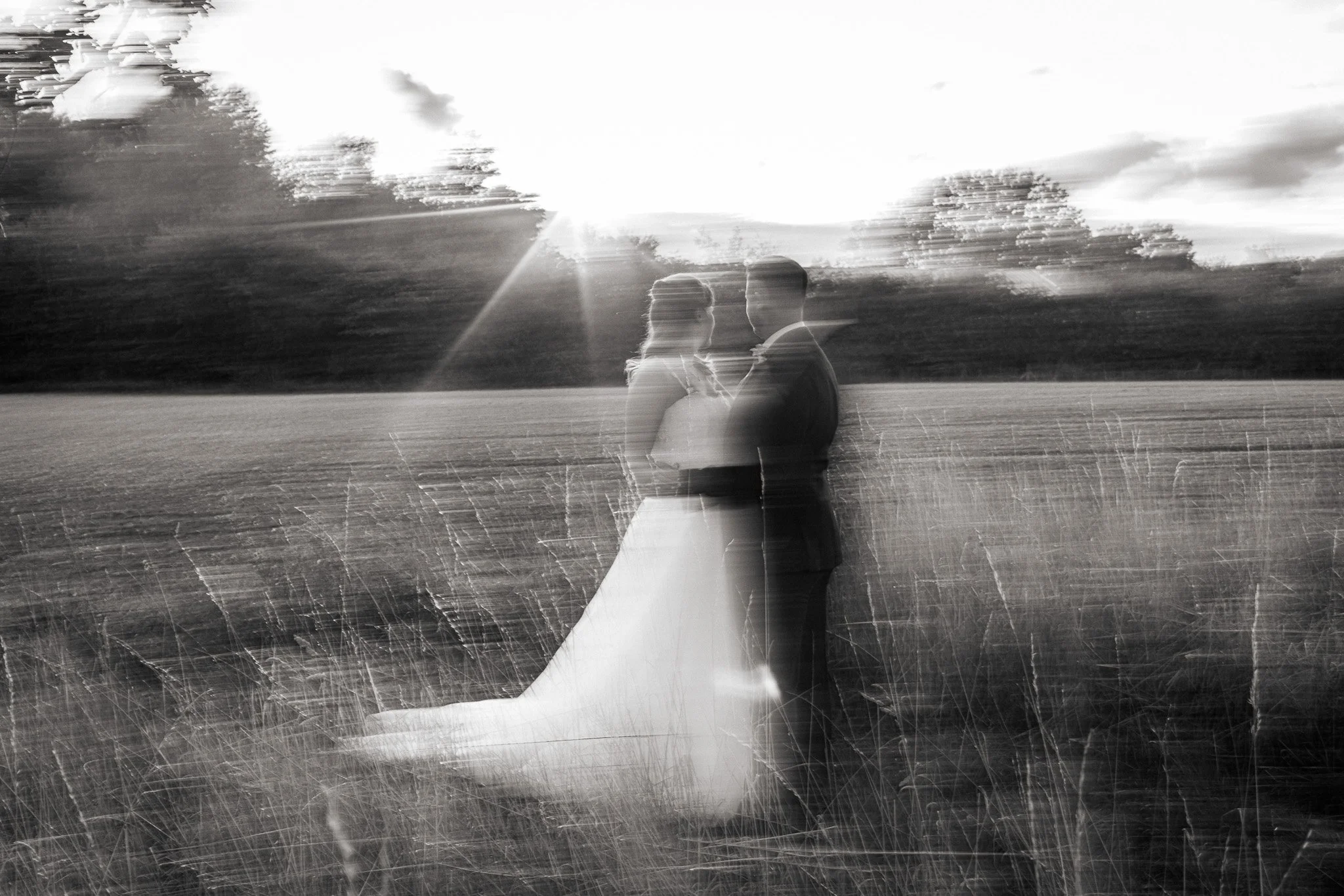 A black-and-white photograph of a couple walking outdoors, with the sun setting or rising behind them, creating a silhouette and lens flare effect, with trees and open field in the background.