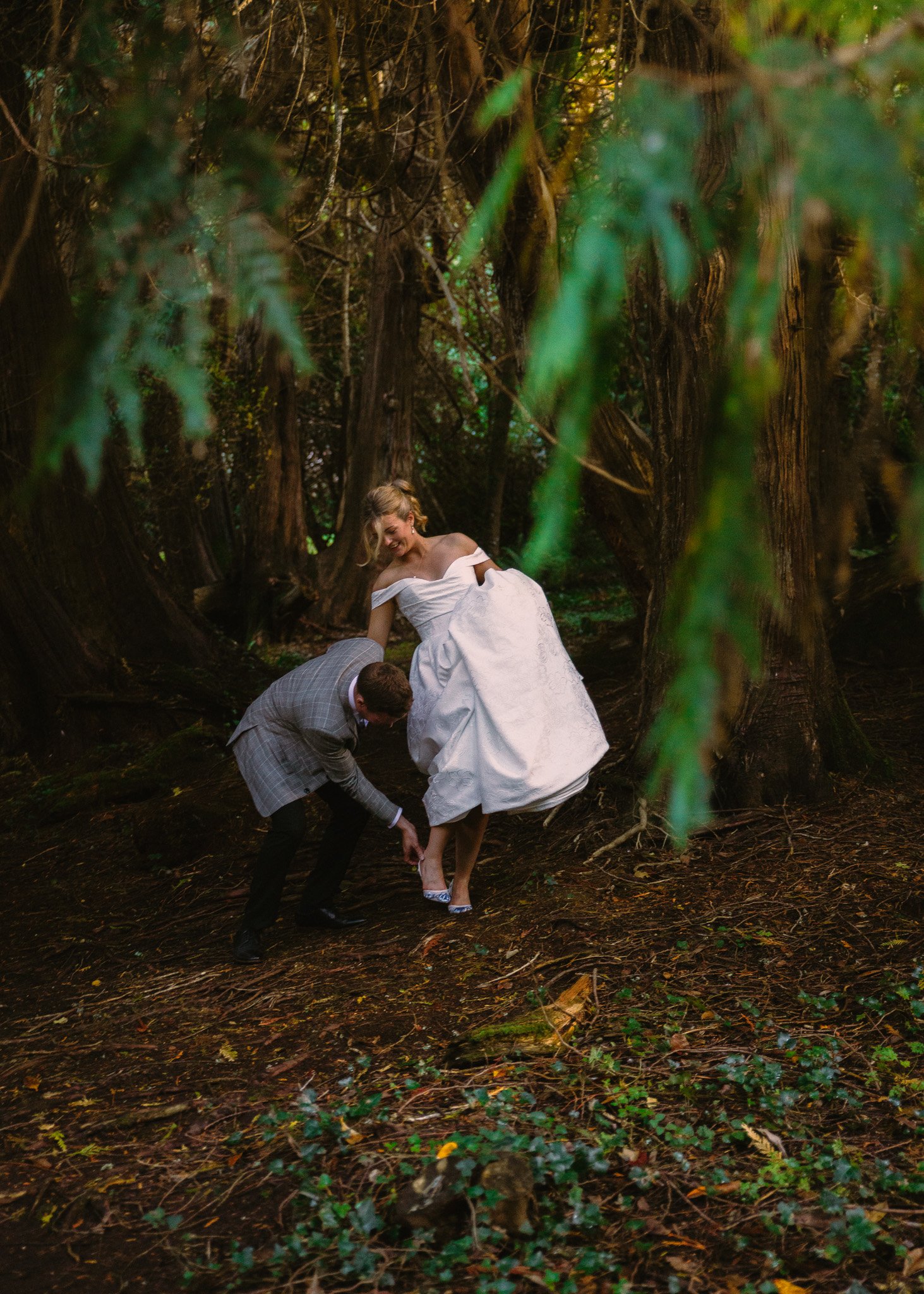 A woman in a white dress poses while a man in a suit helps her with her shoe in a wooded area.