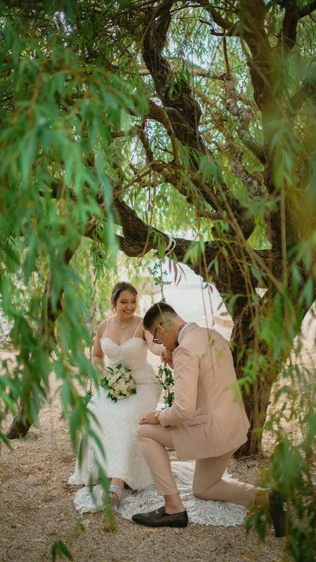 A couple in wedding attire under a large leafy tree, with the groom kneeling and proposing to the bride, who is smiling and holding a bouquet of flowers.
