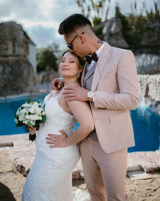 A newlywed couple standing outdoors in front of a pool, with the groom kissing the bride's forehead. The bride is holding a bouquet of white flowers, wearing a white lace wedding dress, and the groom is dressed in a beige suit with a bow tie and glasses.