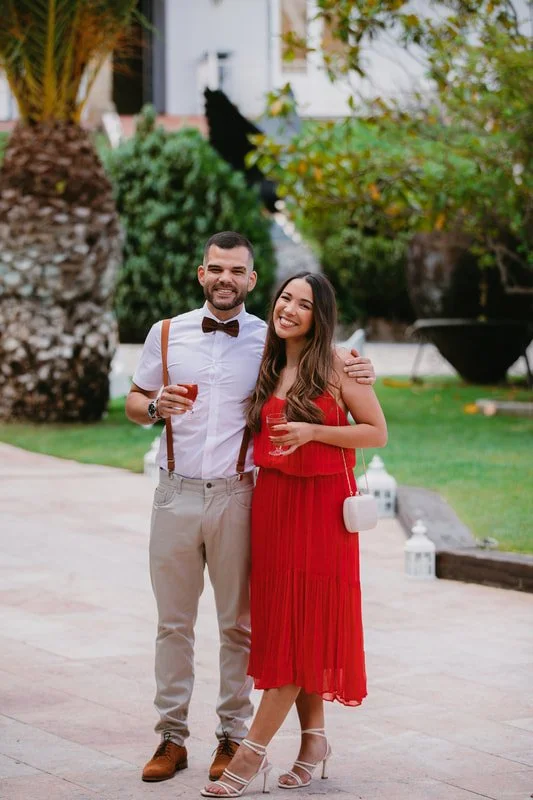 A man and woman dressed formally, smiling and holding drinks, standing outdoors in a garden area with trees and plants in the background.