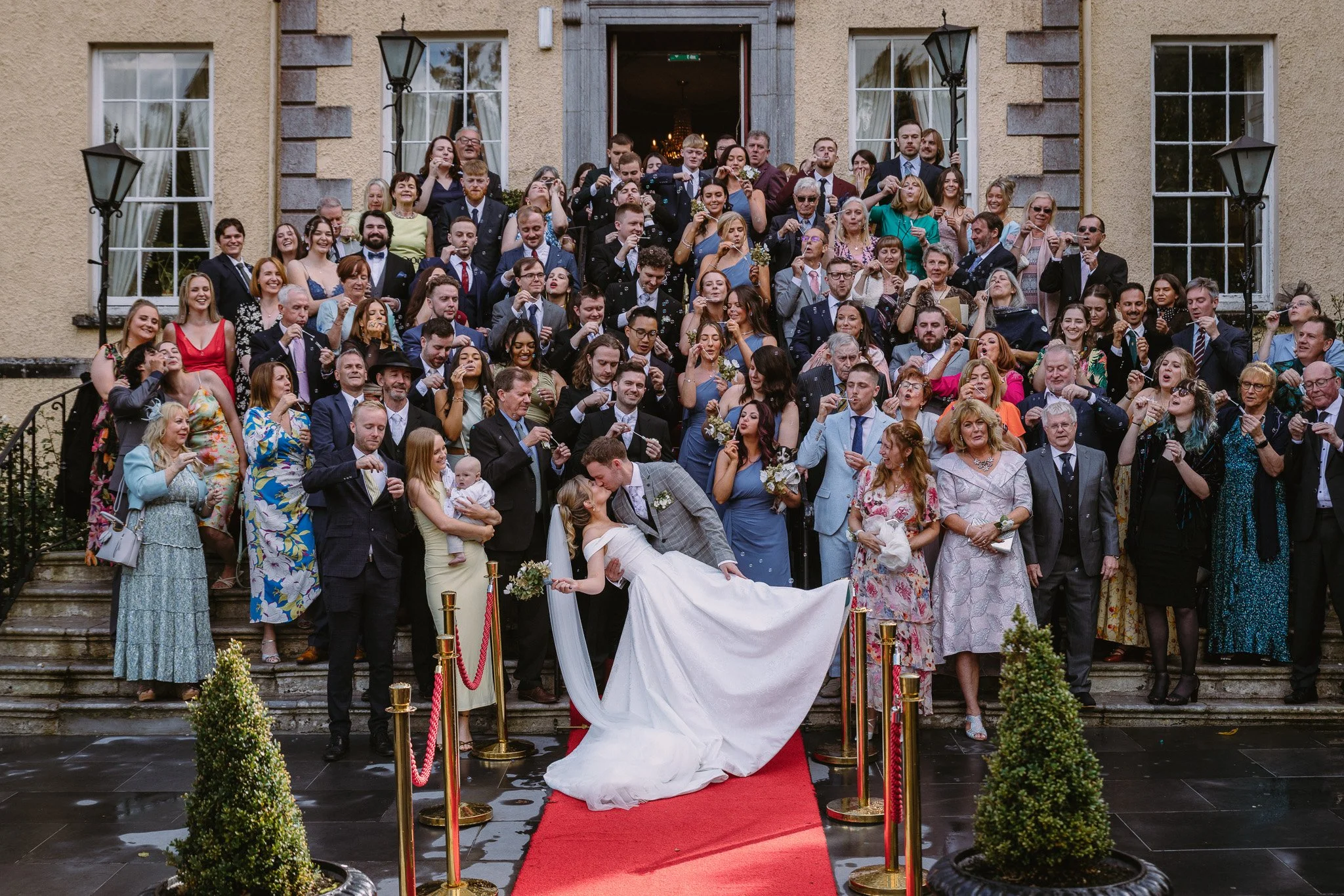 A large group of people, including men, women, and children, gathered on the steps of a building for a wedding celebration. The bride and groom are at the front, with the bride holding the groom's head and a man lifting her in a embrace. Everyone is dressed in formal attire.