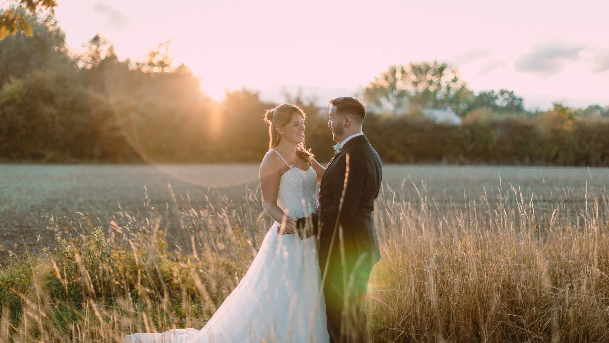 A bride and groom standing in a field at sunset, holding hands and gazing at each other with trees in the background.