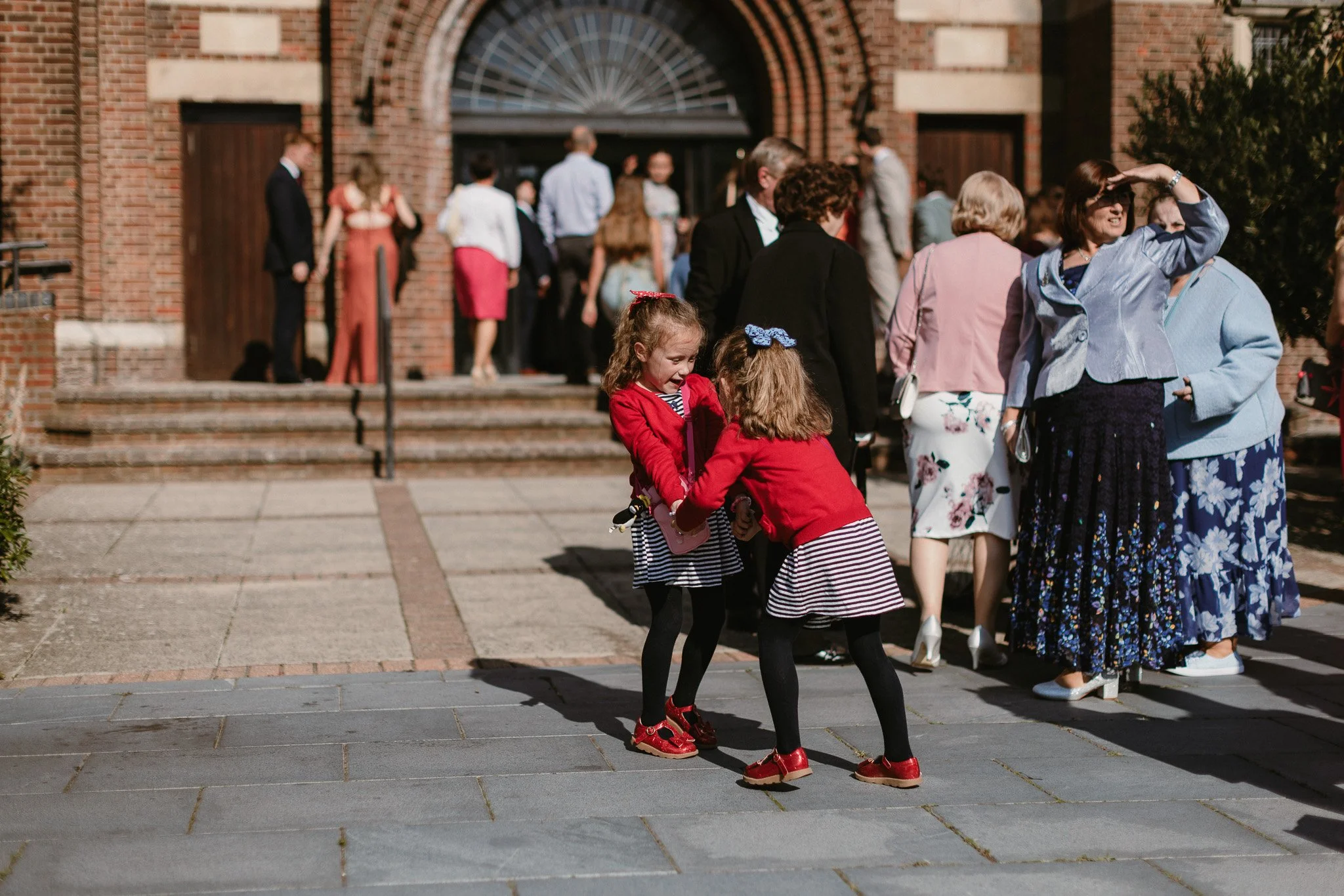 Two young girls in matching striped skirts and red jackets are laughing and holding hands in front of a group of adults outside a brick building, with a set of steps and large wooden doors in the background.