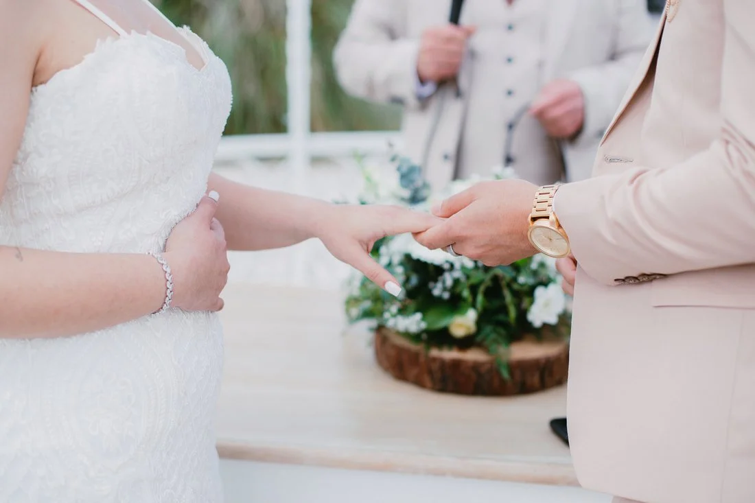 A wedding ceremony where a bride in a white lace dress and a groom in a light beige suit are exchanging rings.