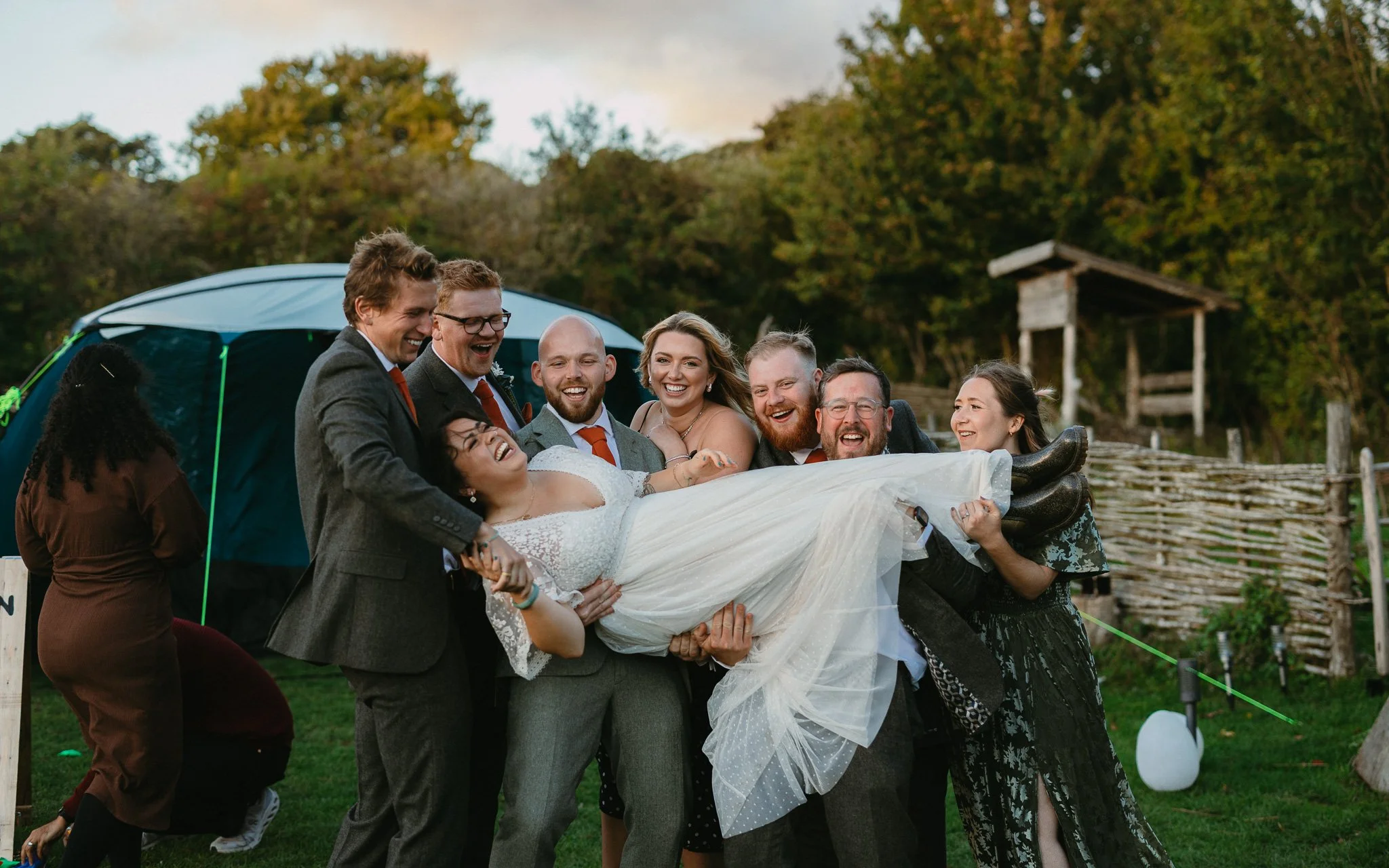 Group of people lifting a woman in a wedding dress outdoors during daytime, with trees and a wooden fence in the background.