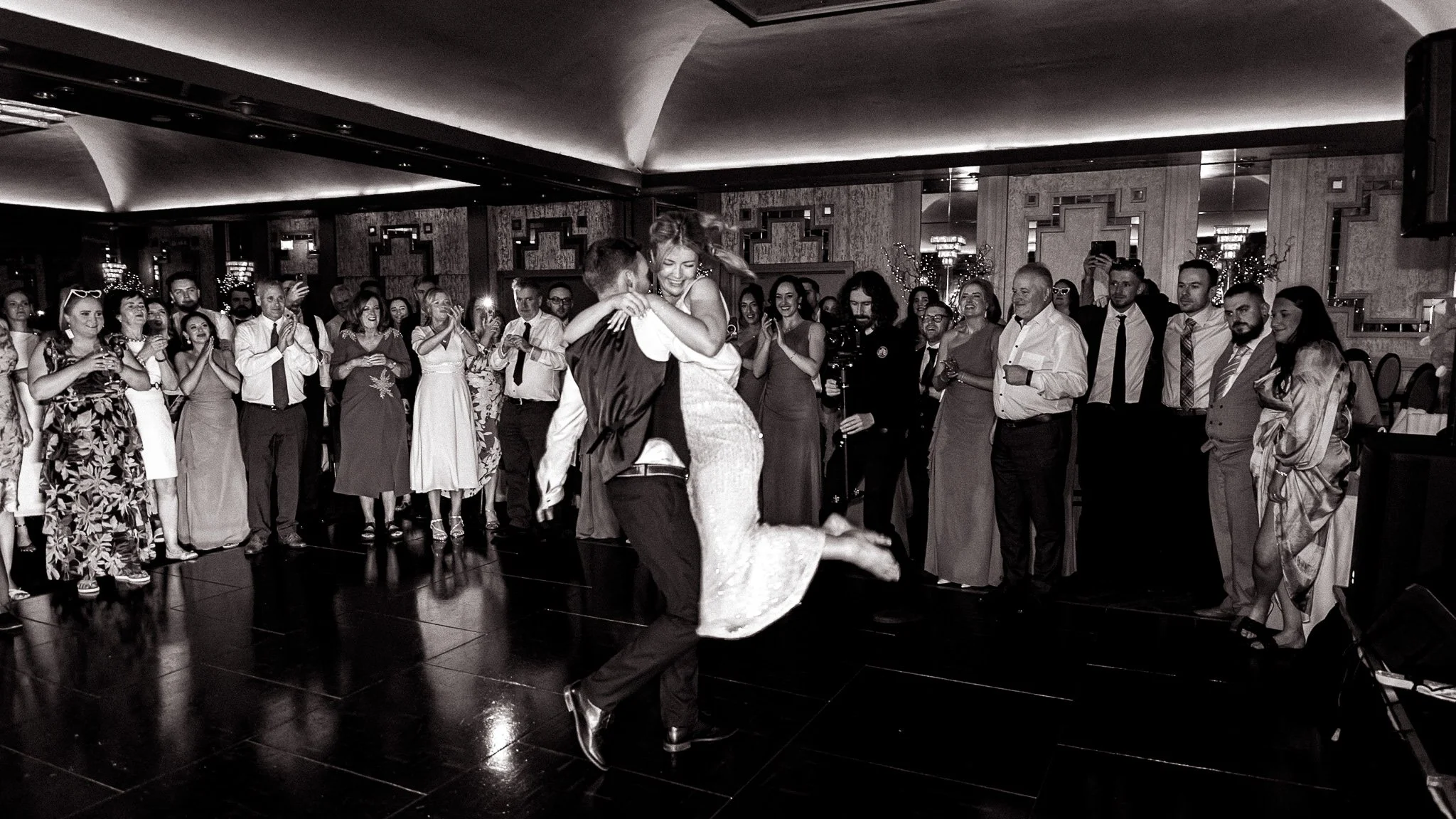 A bride and groom dancing at their wedding reception, surrounded by guests clapping and watching in a decorated banquet hall.