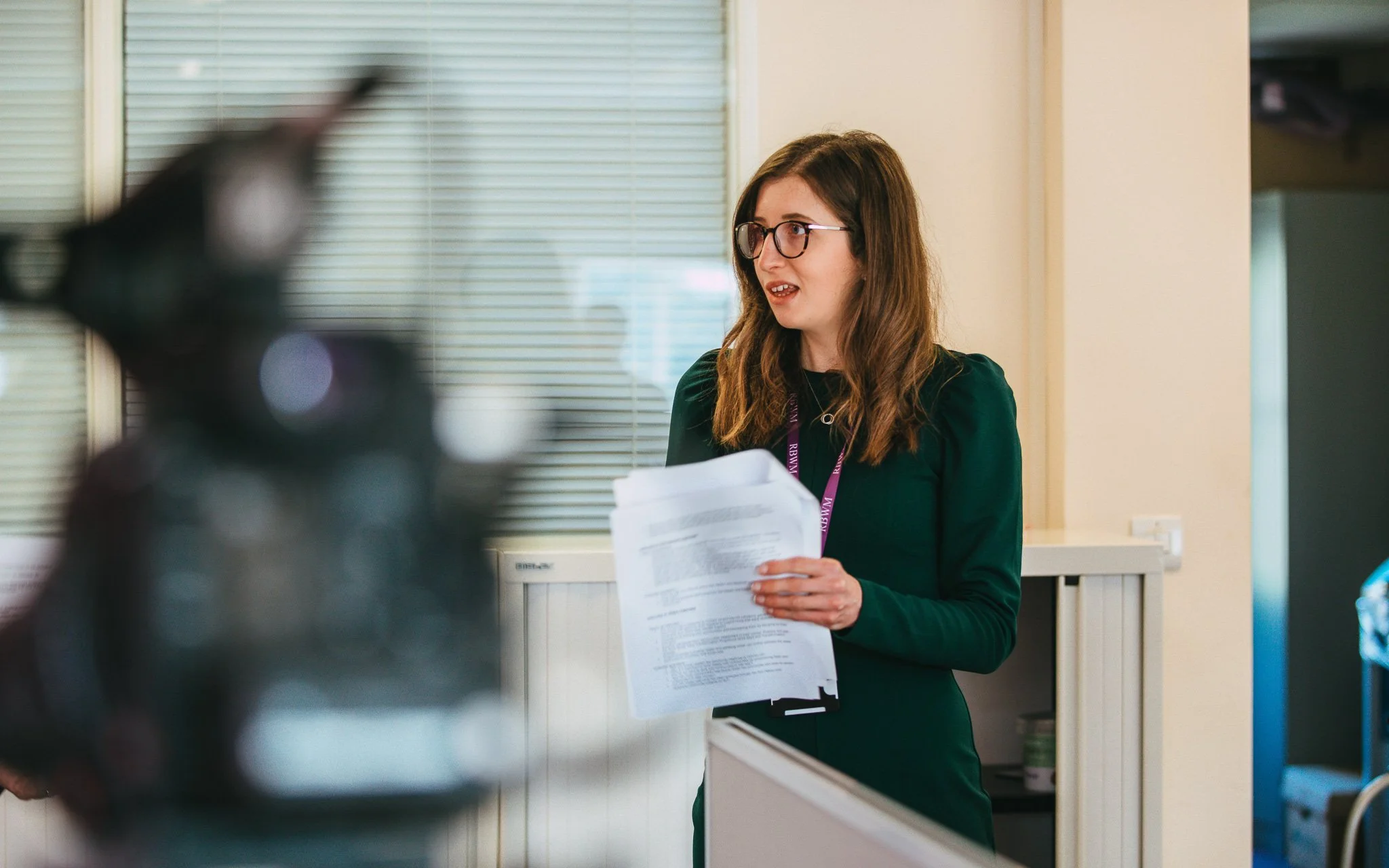 A woman with glasses holding a stack of papers in an office setting.