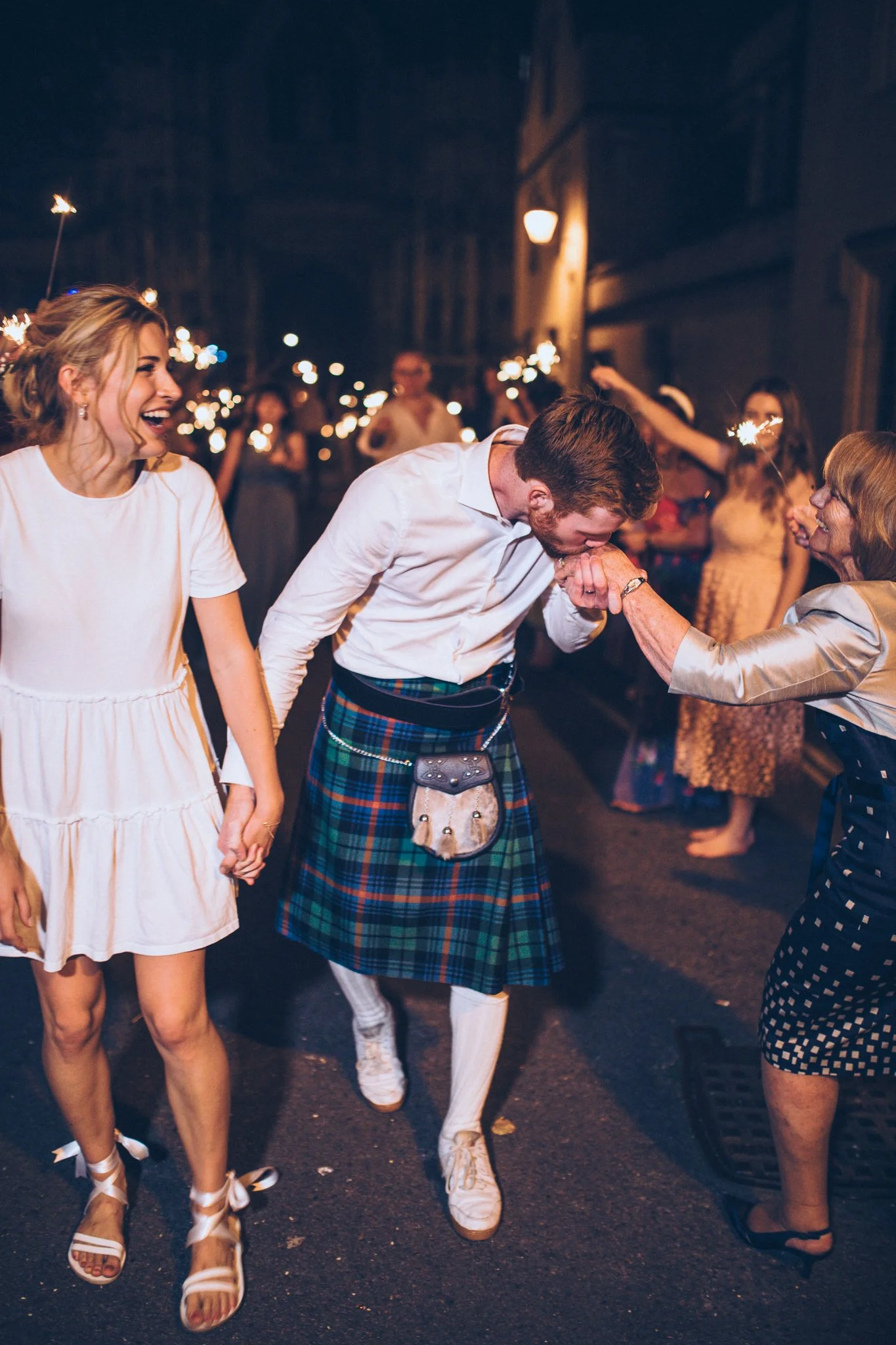 People celebrating at night with sparklers, including a woman in a white dress and sandals, a man in traditional Scottish attire, and others in the background holding sparklers.