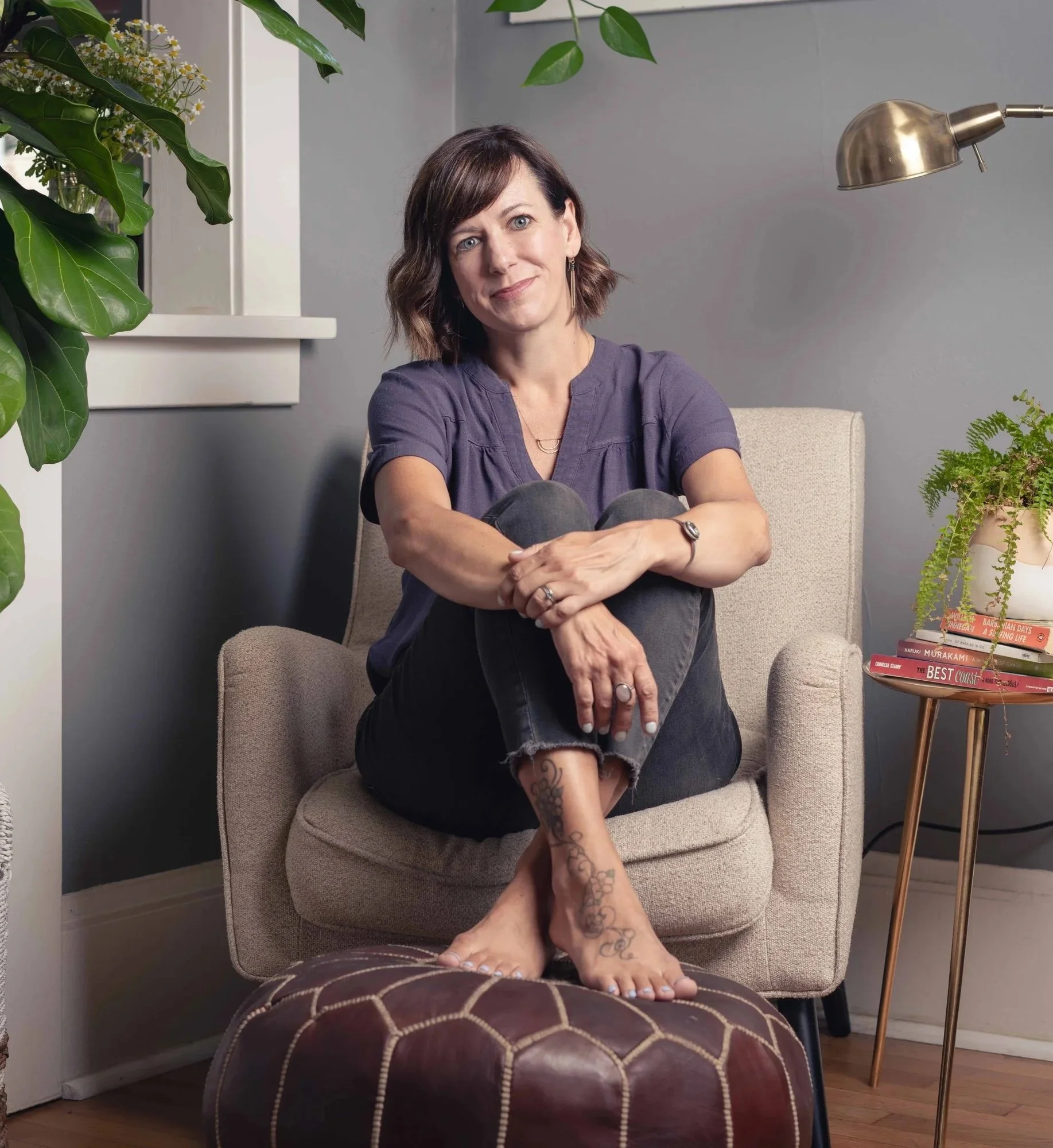 A woman with short brown hair, blue eyes, and earrings sitting on a beige armchair with her legs crossed and her arms resting on her knees. She is smiling slightly and looking at the camera. She is wearing a purple shirt and has tattoos on her left ankle and foot. Behind her, there are green plants, a gray wall, a stack of books, and a floor lamp.