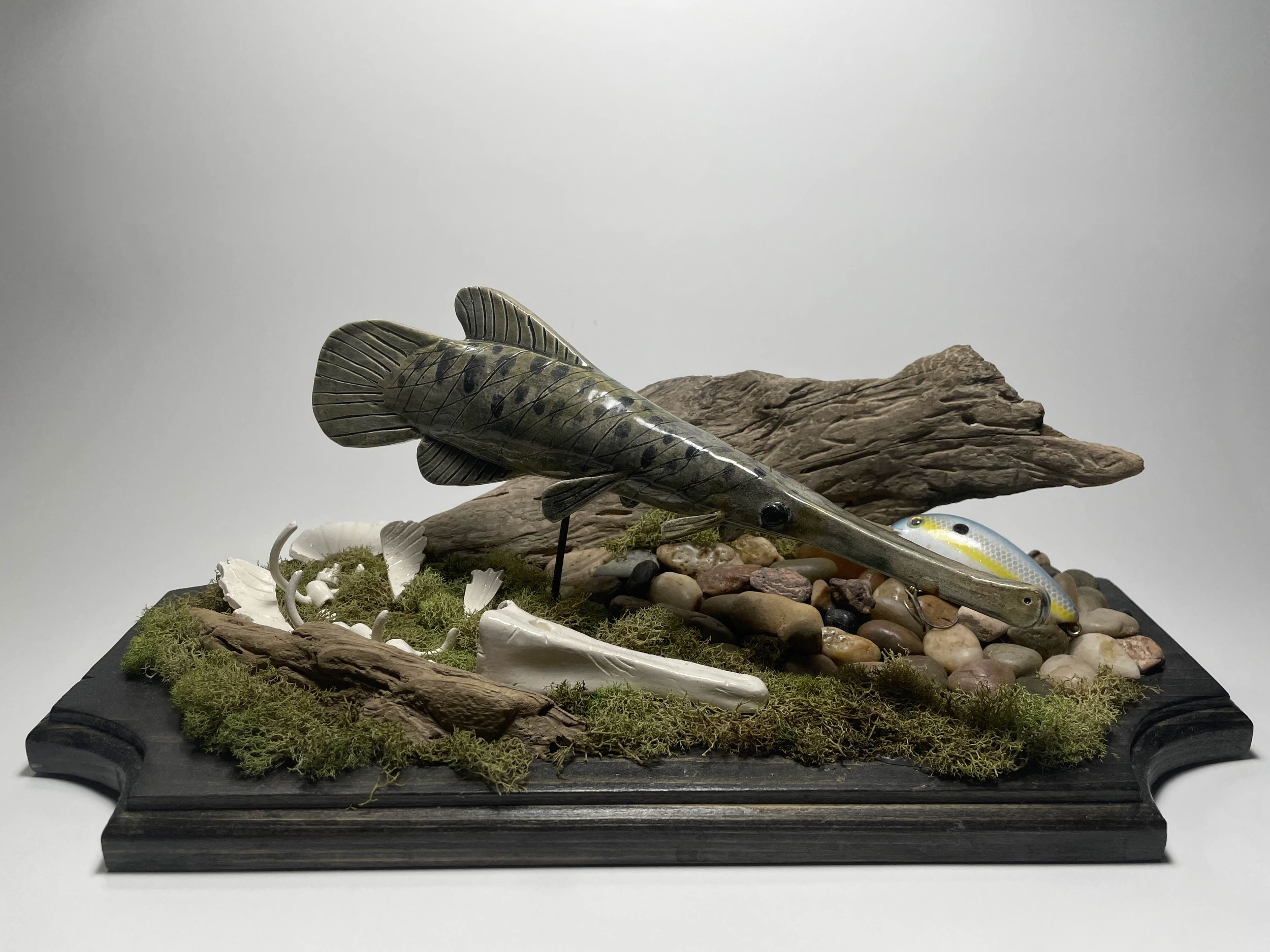 Decorative fishing-themed display with a fish sculpture, rocks, driftwood, and moss on a wooden base.