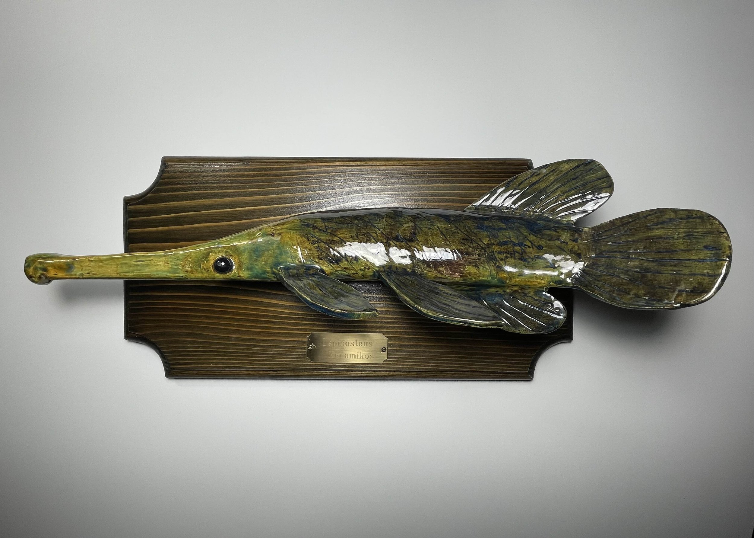 A decorative wall plaque holding a fish with a gold and greenish glaze, mounted on a wooden background with a metal plaque that reads "Lepestoseus Keramicus".