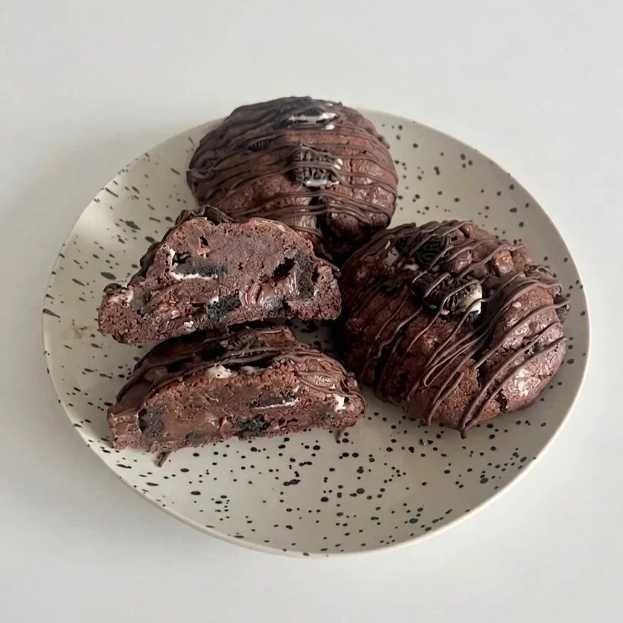 Four chocolate cookies drizzled with chocolate on a white speckled plate, with one cookie cut in half showing chocolate chips inside.