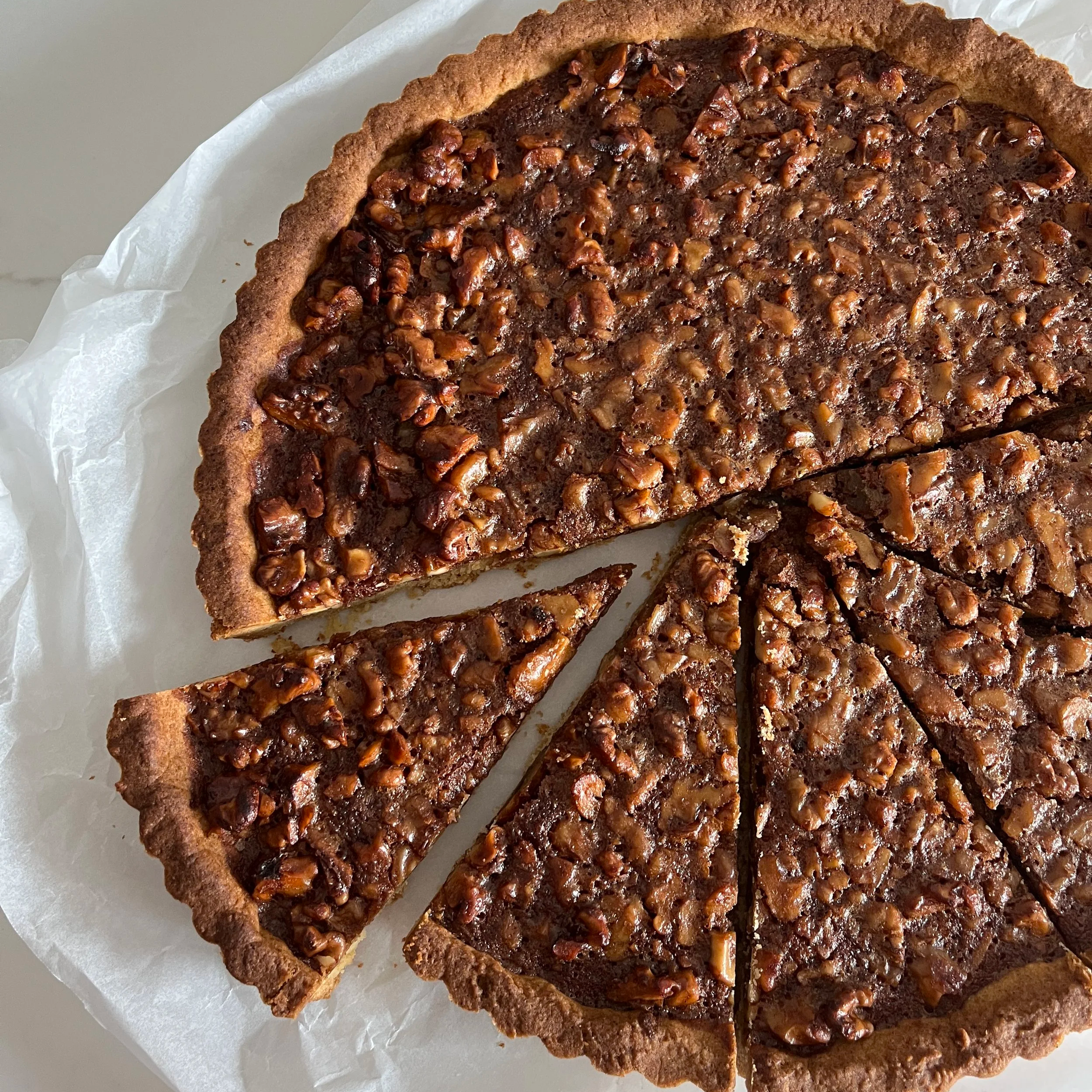 Chocolate pecan tart with a golden crust and chopped pecans on top, sliced on white parchment paper.