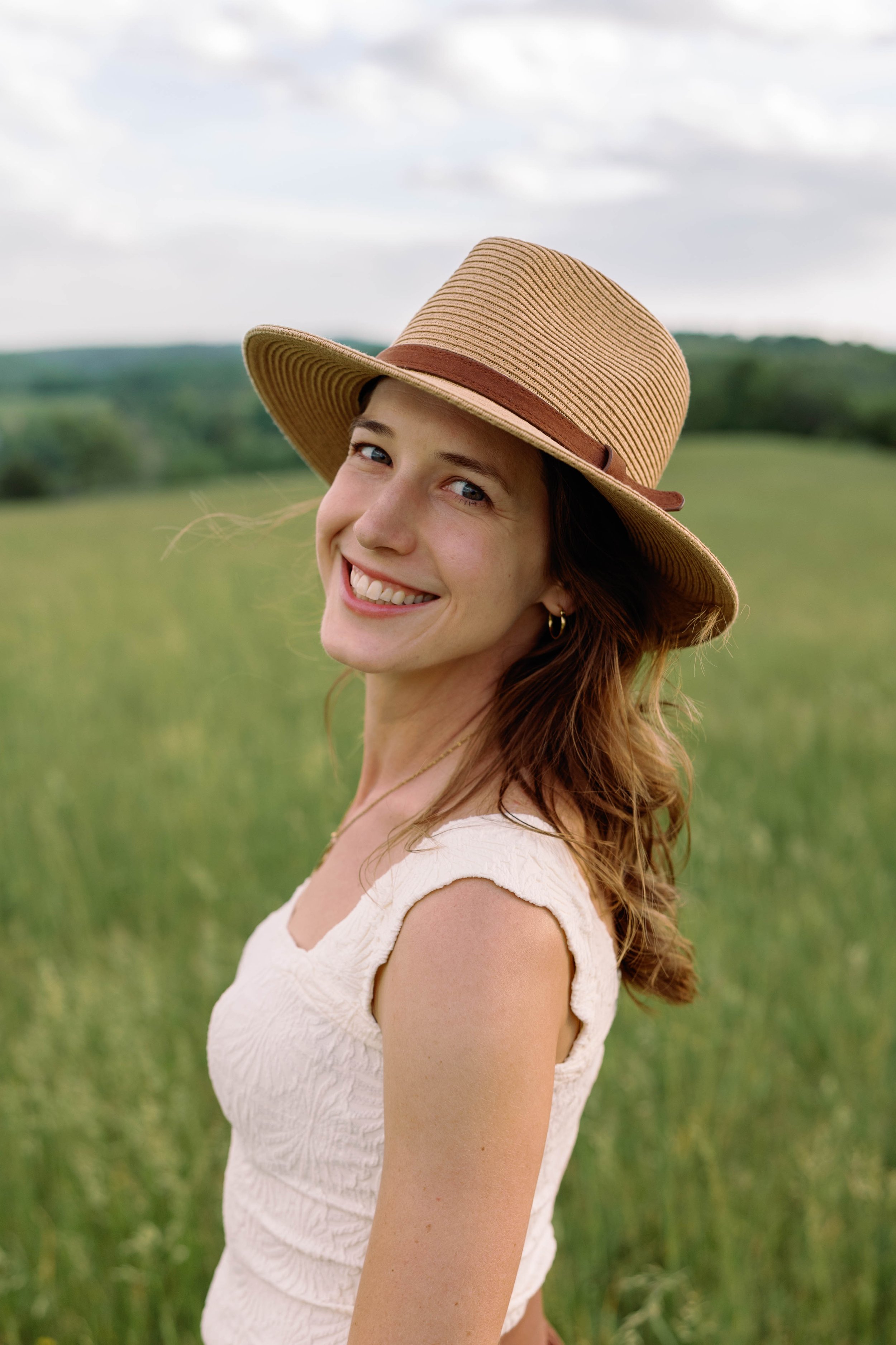 Contact Jessie at Align Integrative Health. Happy woman with red hair wearing a tan wide-brimmed hat and a light-colored sleeveless dress, standing in a green field with rolling hills and cloudy sky in the background.