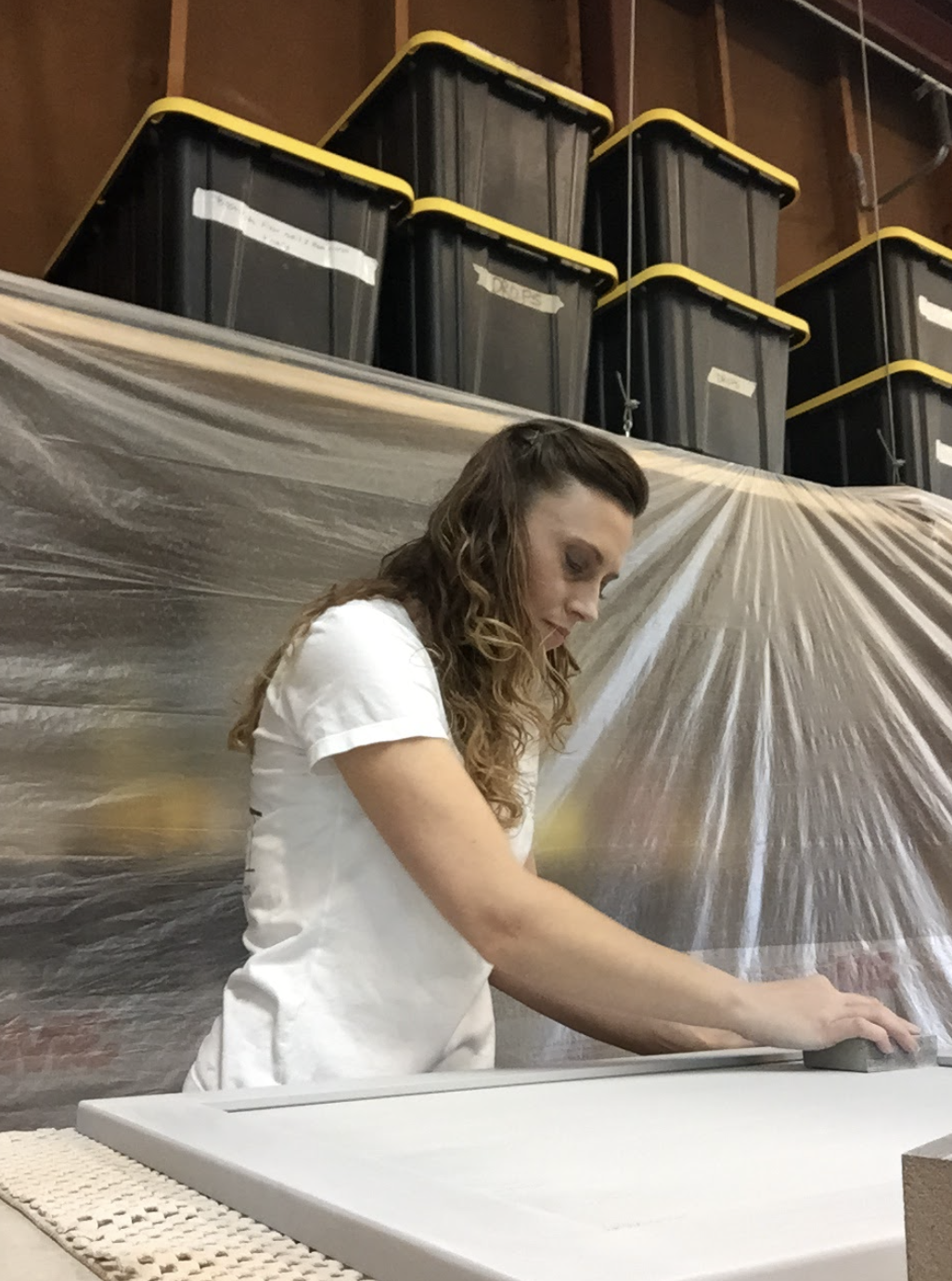 A woman wearing a white shirt is working on a large flat surface, possibly in a workshop or studio. She is focused on her task, with her right hand holding an object or tool. Behind her, there is plastic sheeting and several black storage containers with yellow lids stacked on a shelf.