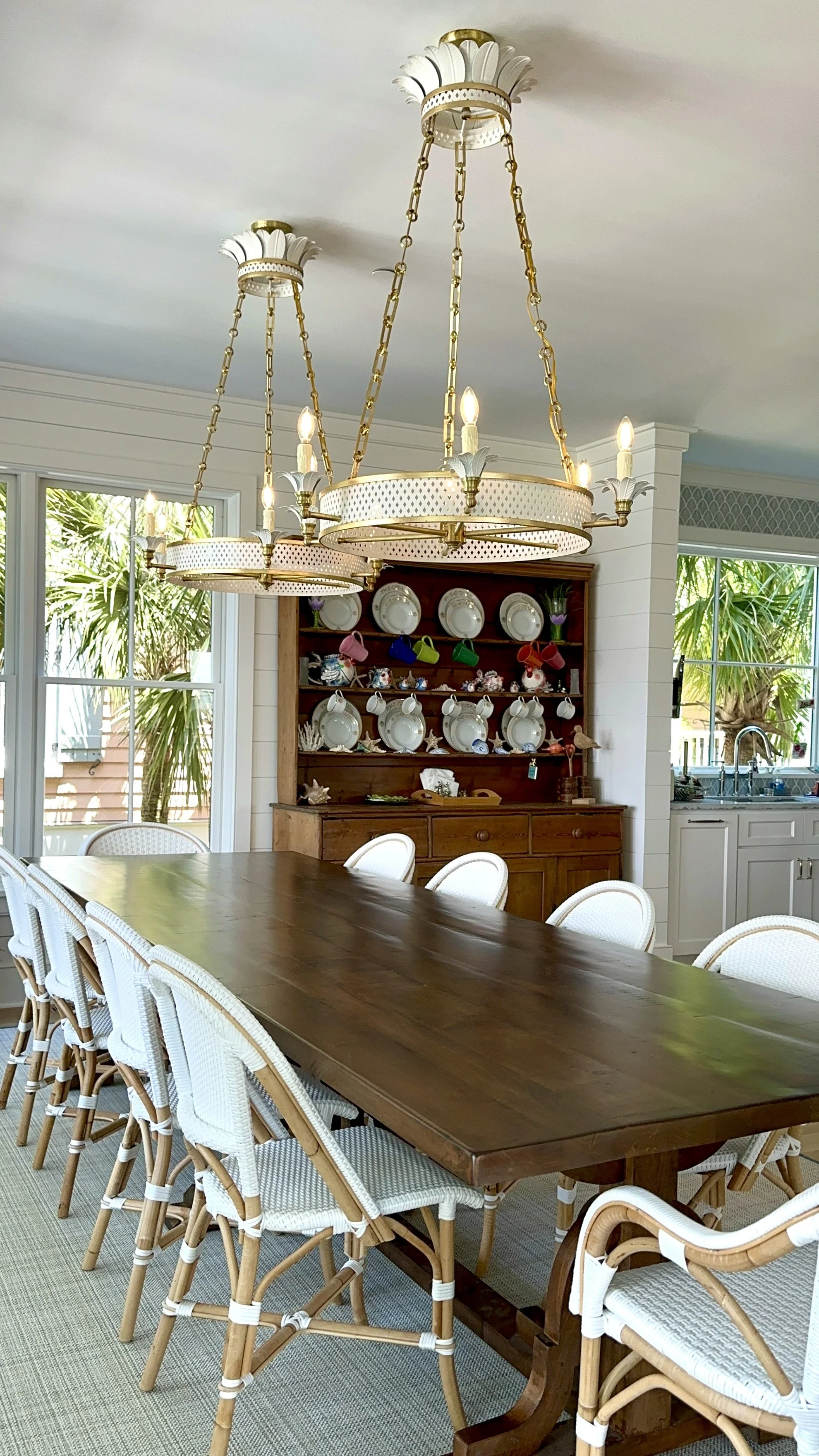 A dining room with a large wooden table surrounded by white wicker chairs, two ornate chandeliers hanging from the ceiling, a wooden hutch with white plates, cups, and decorative items, large windows with views of greenery outside, and a kitchen area