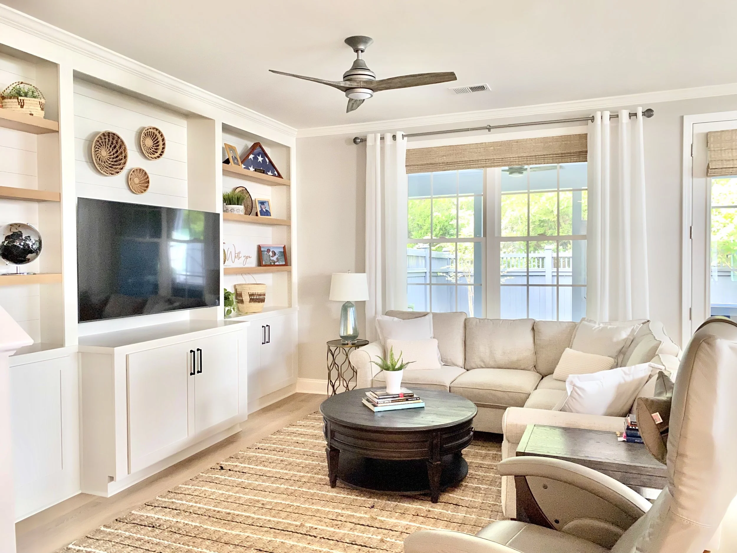 Living room with beige sectional sofa, black coffee table with books, white built-in shelves with decor, flat-screen TV, window with white curtains, ceiling fan with wooden blades, and a patterned area rug.