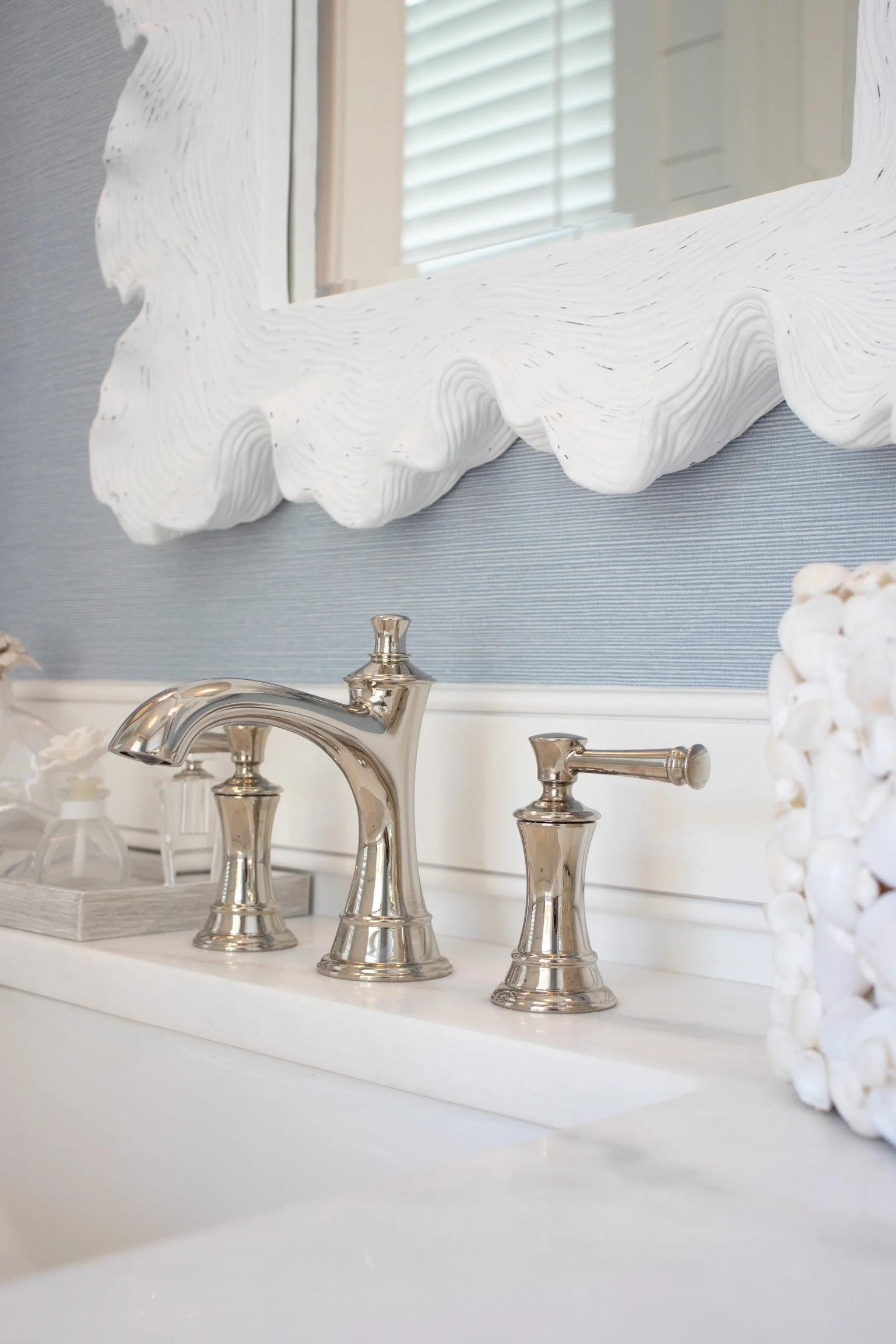 Decorative bathroom mirror with a white, textured, wavy frame above a white bathroom sink with silver fixtures. A window with blinds reflects in the mirror.