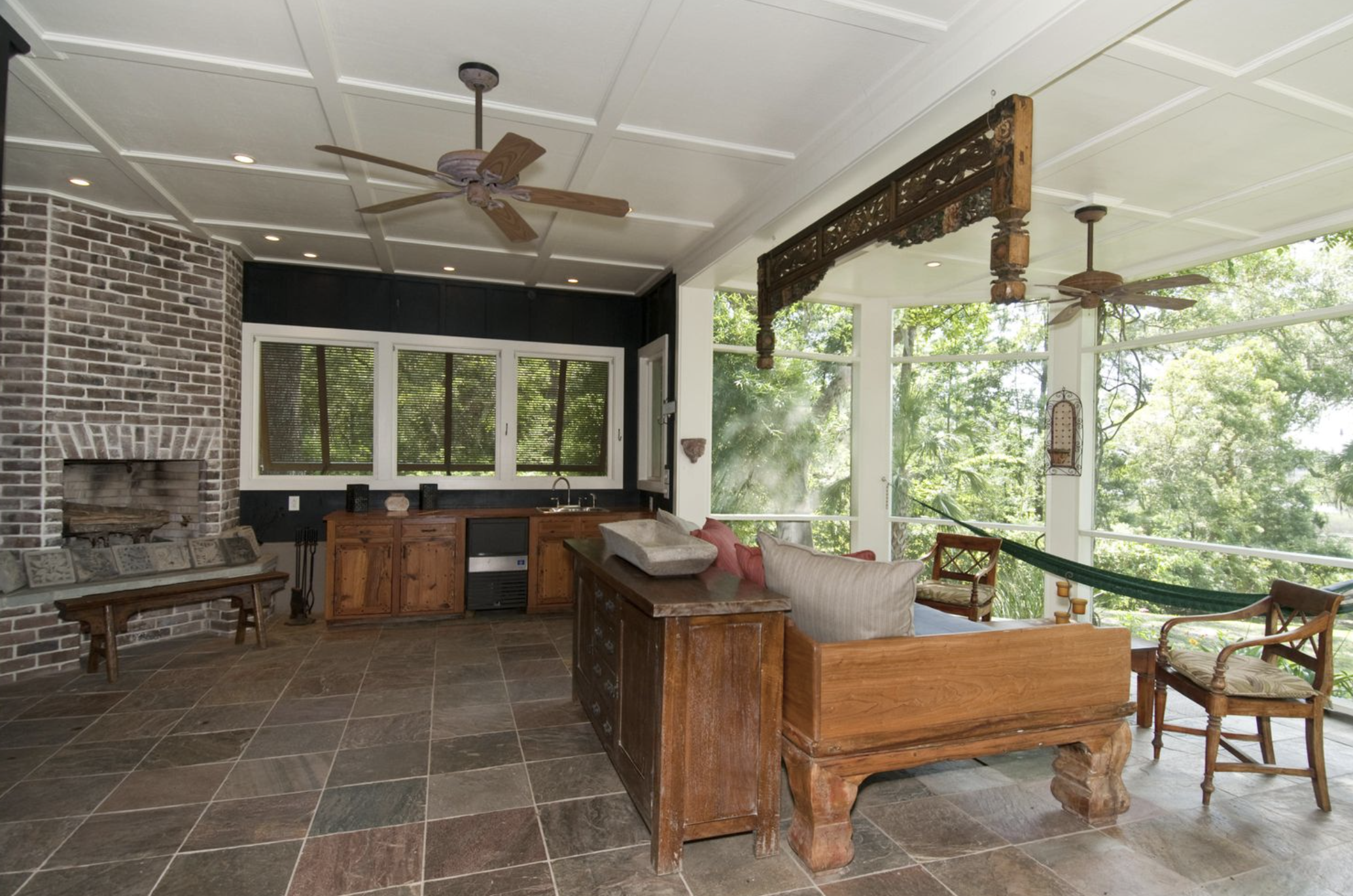 Living room with brick fireplace, wooden furniture, large windows with trees outside, ceiling fans, and indoor plants.