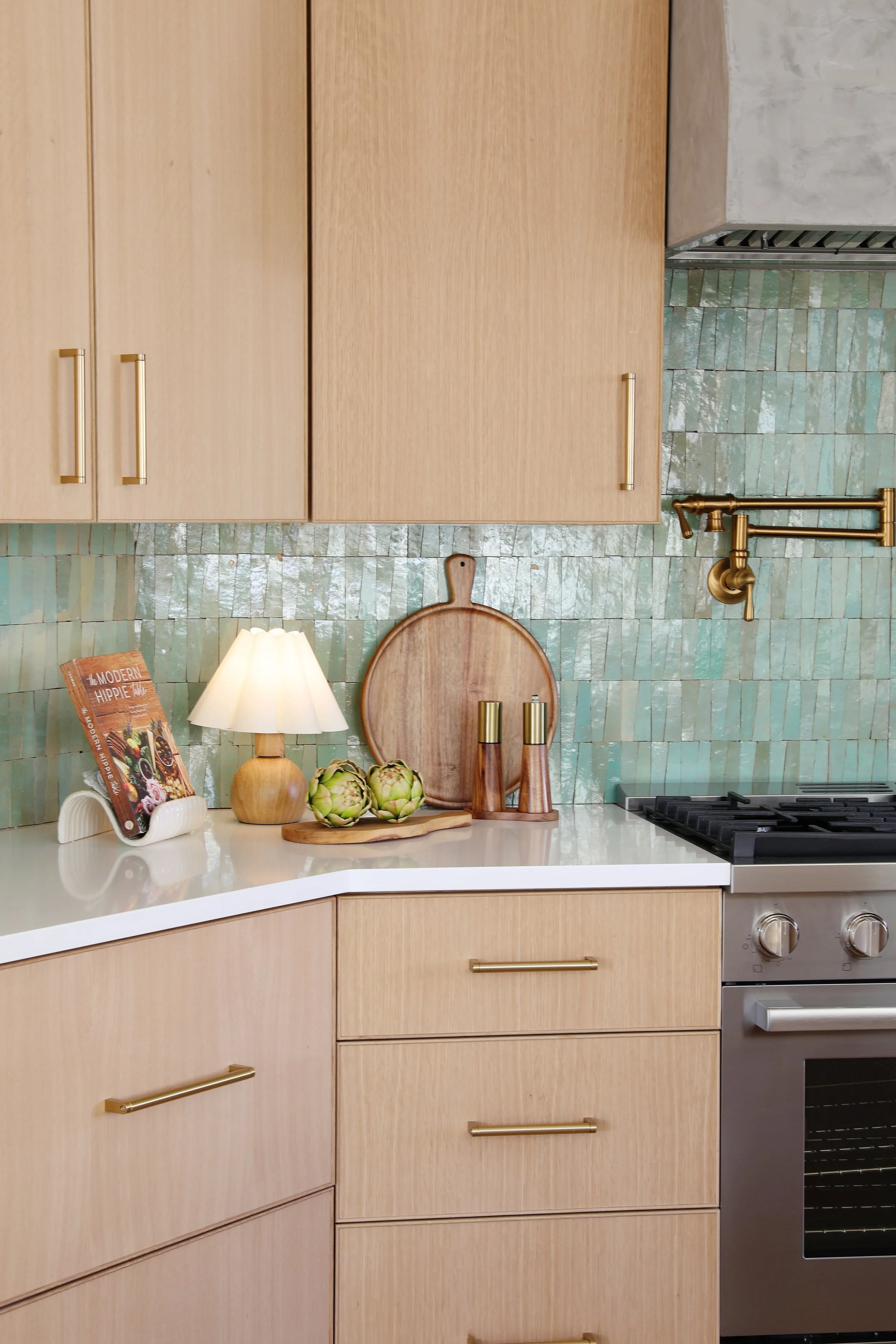 A kitchen with light wooden cabinets, green tiled backsplash, and a stainless steel stove. Countertop decor includes a small lamp, artichokes, a wooden cutting board, and brass kitchen utensils.