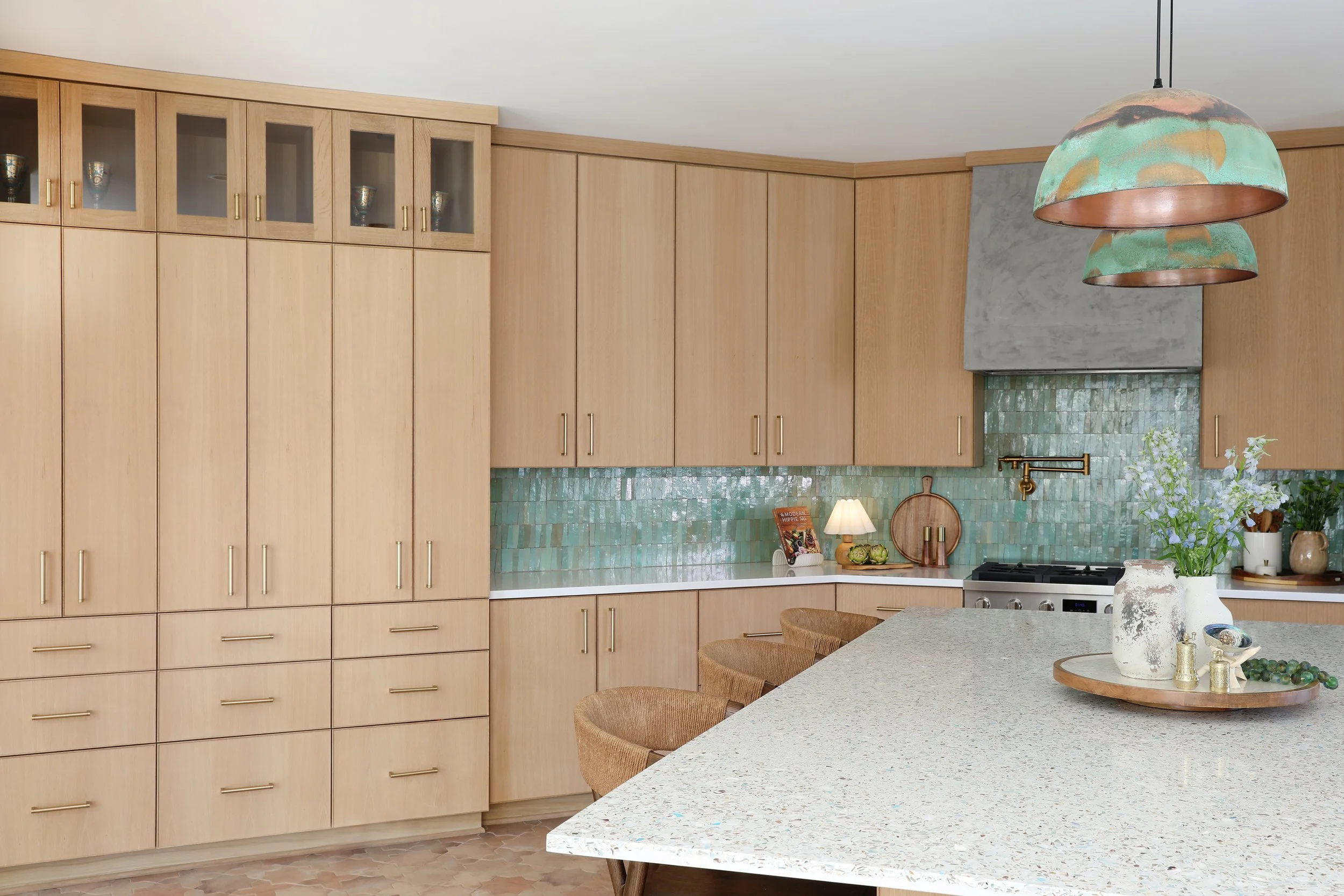 Modern kitchen with light wood cabinets, green mosaic tile backsplash, white countertop, and decorative items on the island including a bowl of grapes and a vase with flowers. A copper and green pendant light hangs from the ceiling.