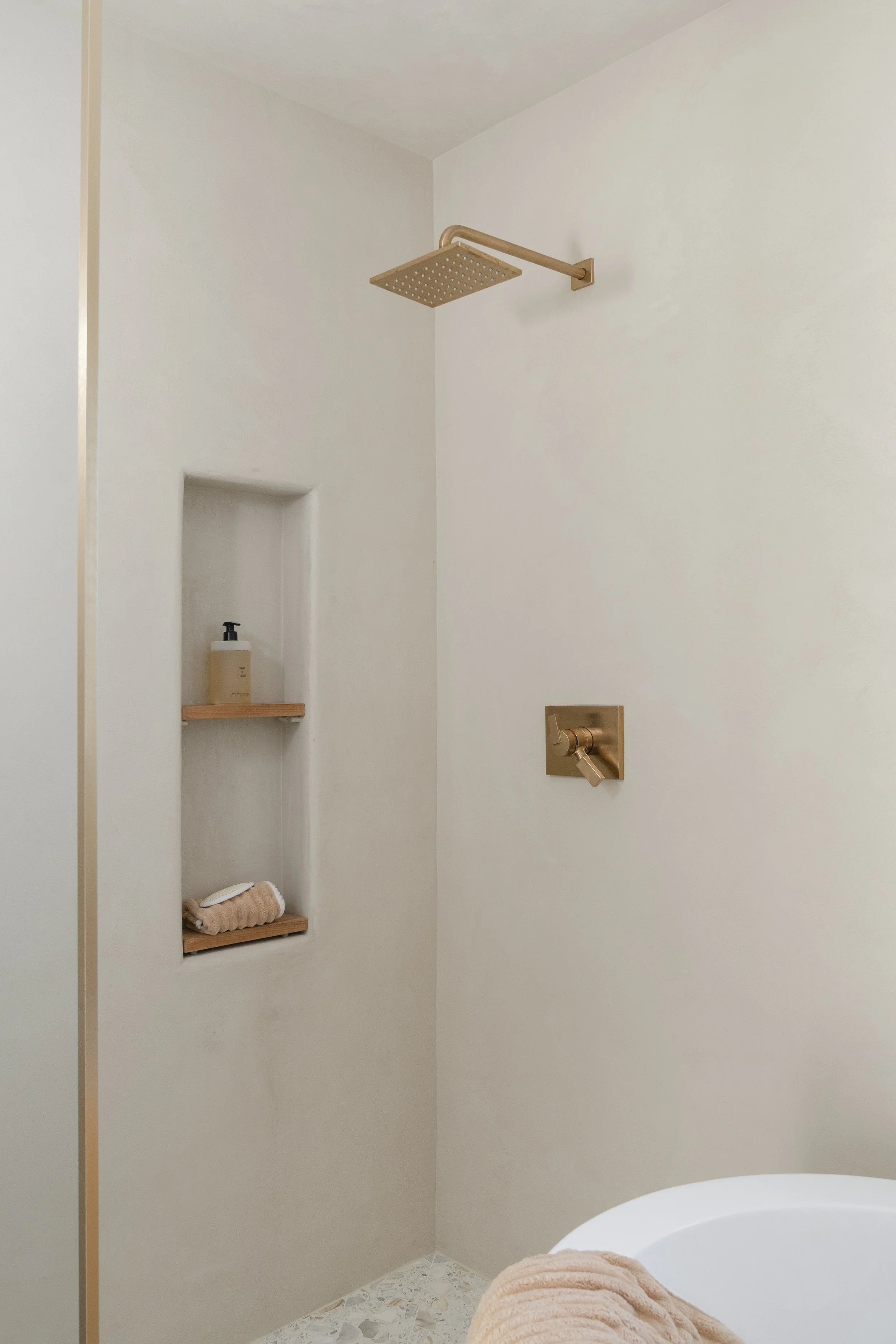 A bathroom shower area with a large, square, gold-colored rainfall showerhead mounted on a beige arm on the wall. There are two built-in shower niches; the upper niche contains a bottle of body wash, and the lower contains a beige towel. Part of a wh