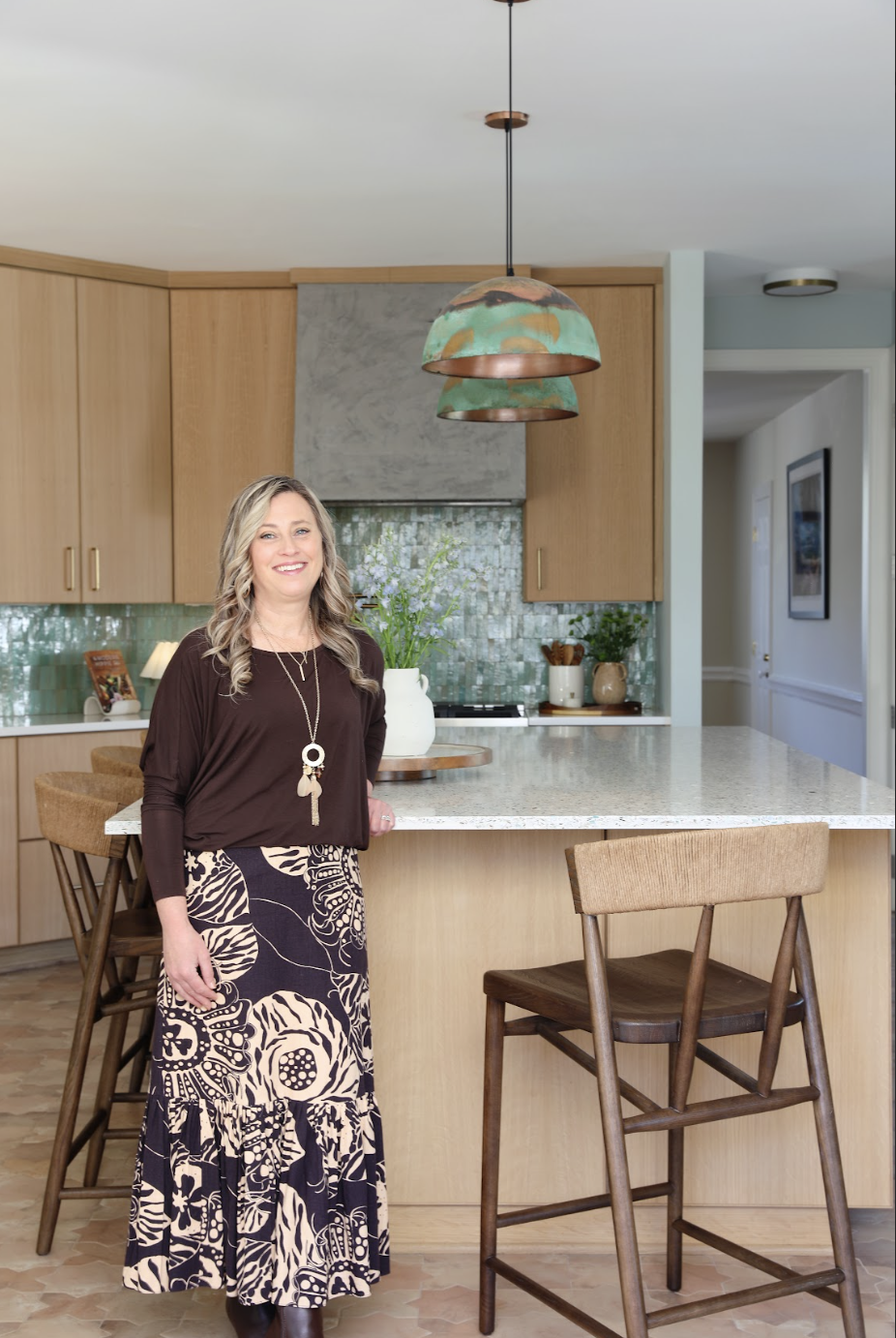 A woman with blonde curly hair smiling in a modern kitchen, wearing a black long-sleeve top and a patterned long skirt. She is standing beside a kitchen island with wooden chairs, above which hang three green and copper pendant lights.