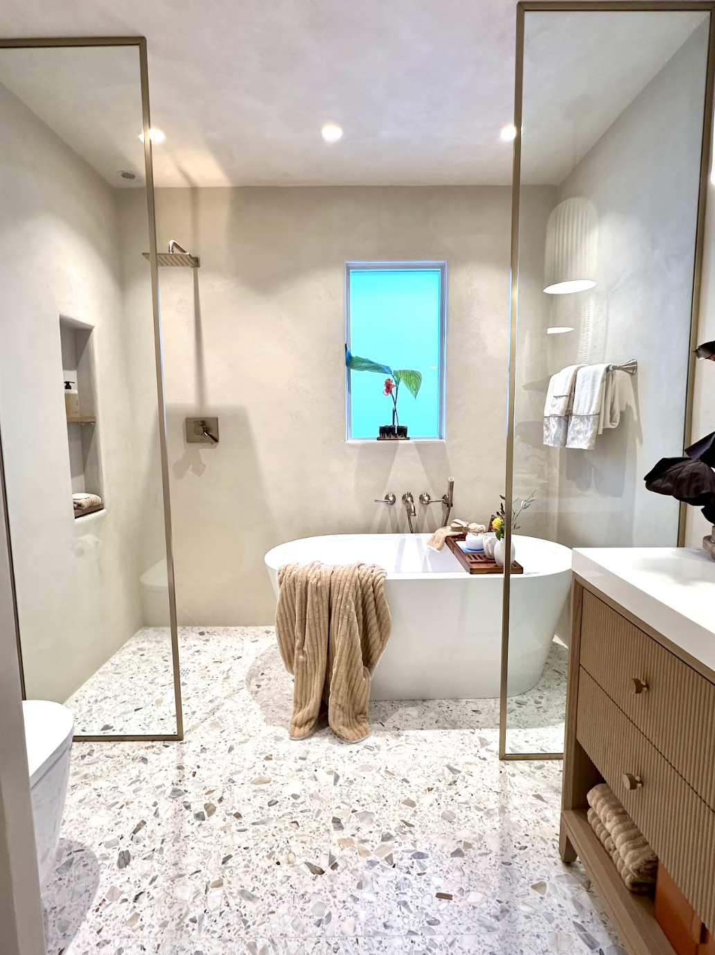 Modern bathroom with a freestanding bathtub beneath a small window, a wood vanity with a towel, a mirror, and a towel rack with towels.