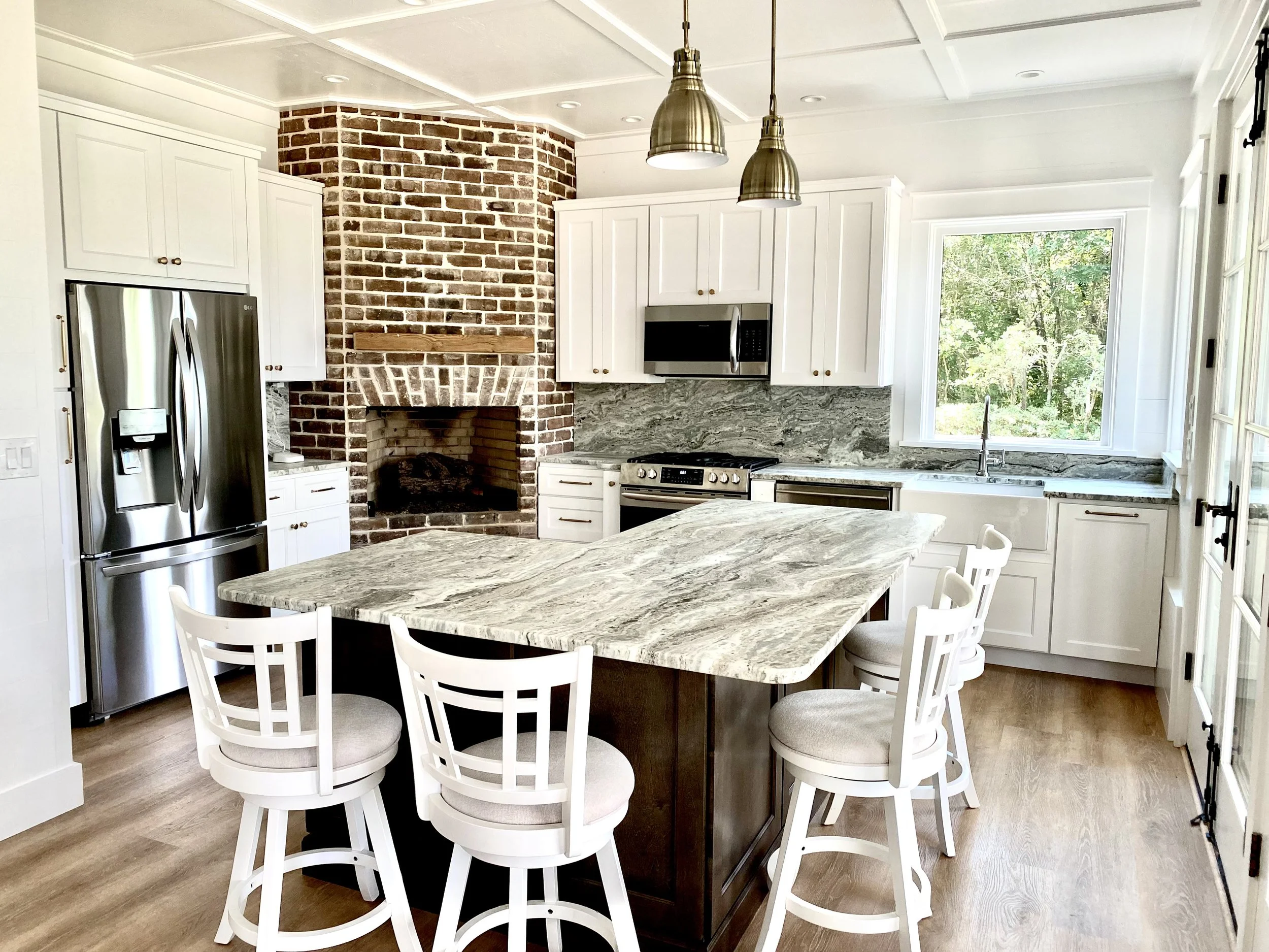 A modern kitchen with white cabinets, a stainless steel refrigerator, a brick fireplace, a marble countertop island, and a large window overlooking greenery.