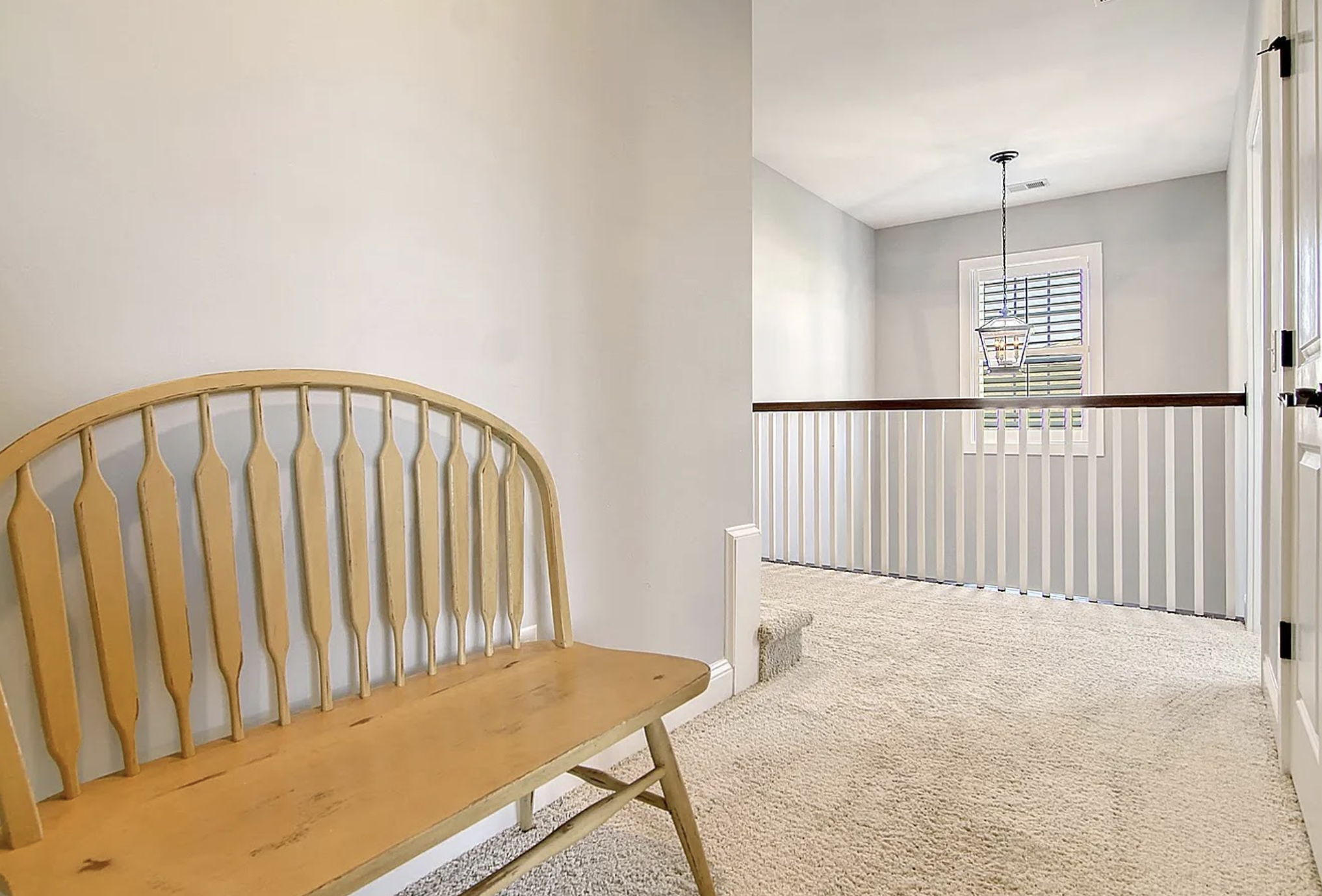 Interior view of a home hallway or landing area with a wooden bench, white railing, window with blinds, and hanging light fixture.