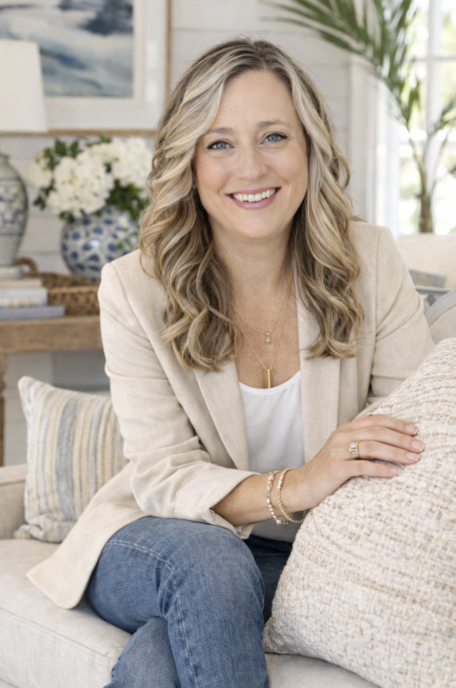 A smiling woman with blonde wavy hair wearing a beige blazer and jeans, sitting on a beige couch in a well-lit living room decorated with plants, flowers, and artwork.