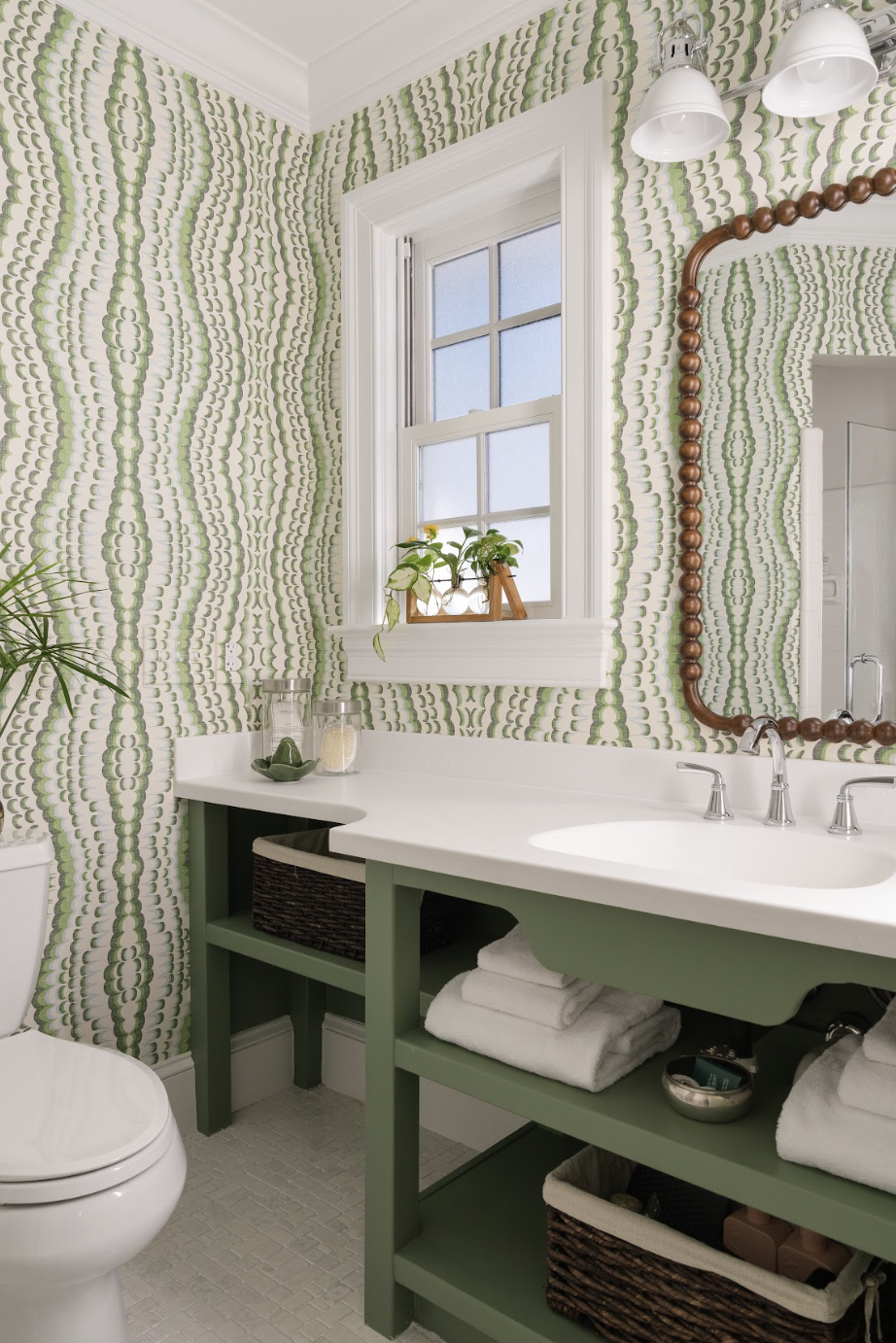 Bathroom with green patterned wallpaper, white vanity with green accents, mirror with wooden frame, window with white trim, plants on windowsill, and bathroom accessories on countertop.