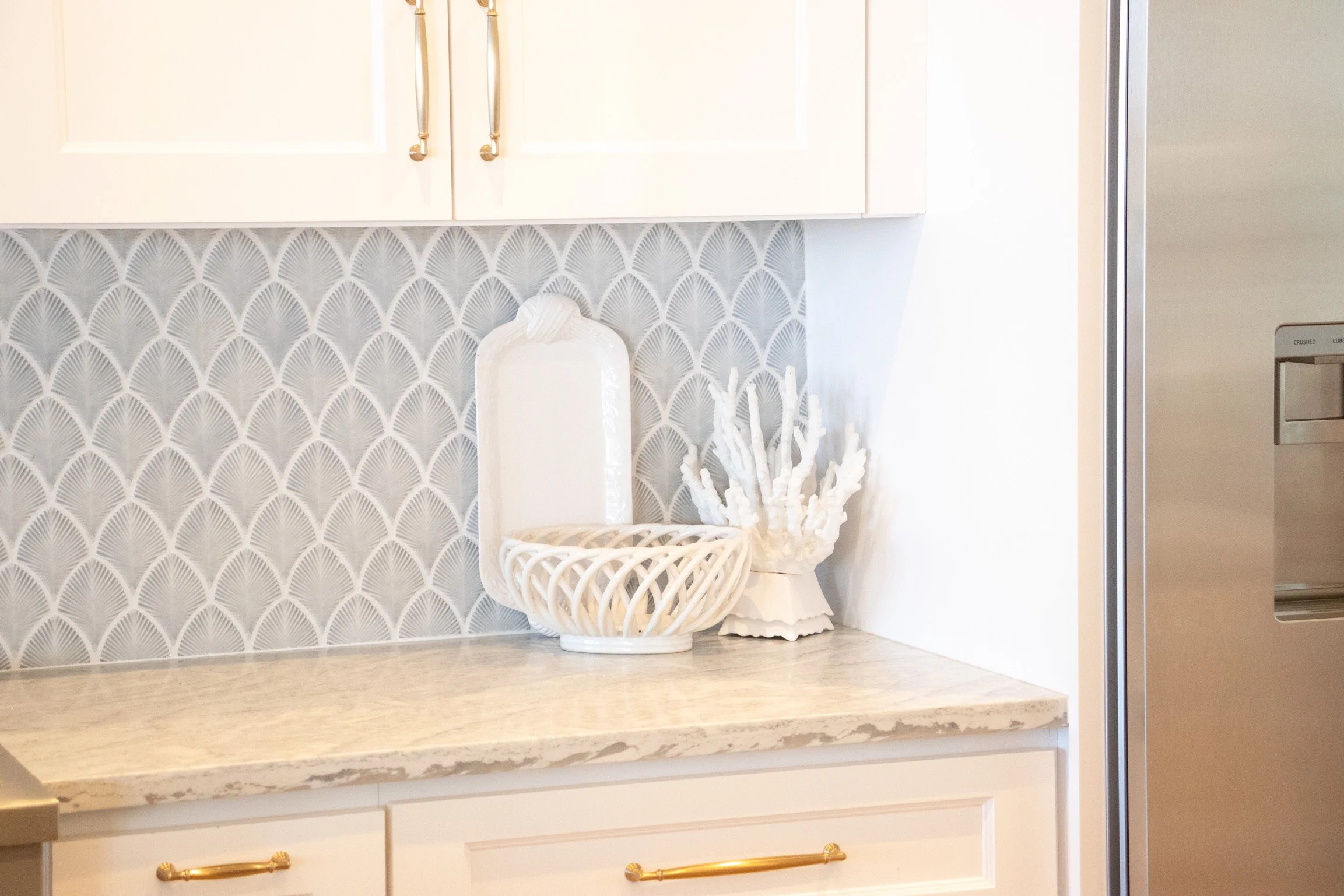 A kitchen countertop with a decorative white vase, a white coral-like sculpture, and a woven basket, with cream cabinets above and a stainless steel refrigerator to the right.