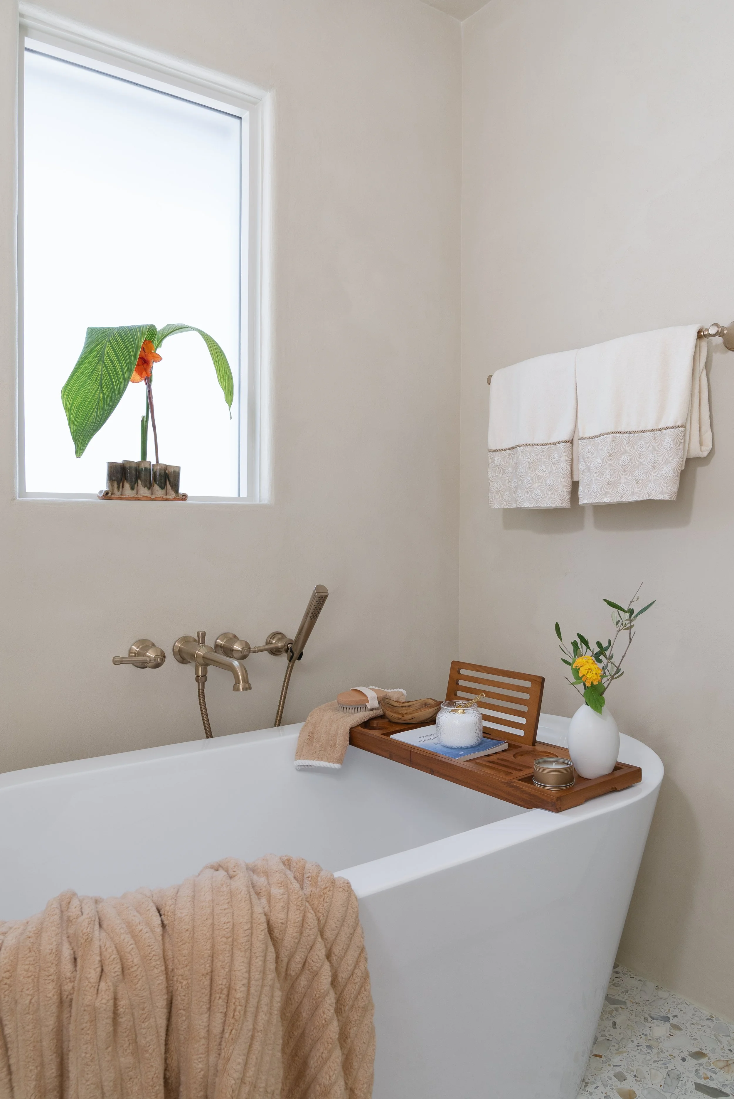 A white bathtub with a wooden tray holding a white vase with yellow flowers, a candle, and a small jar. A beige towel hangs over the side of the bathtub. There is a window with a plant on the windowsill, and a towel rack with a white towel. The floor