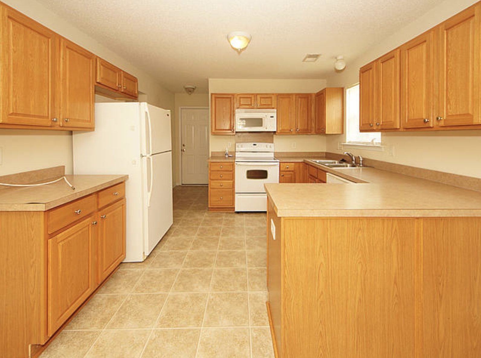 A spacious kitchen with wooden cabinets, beige countertops, and a tiled floor. Includes a white refrigerator, microwave, oven, and sink, with a window above the sink.