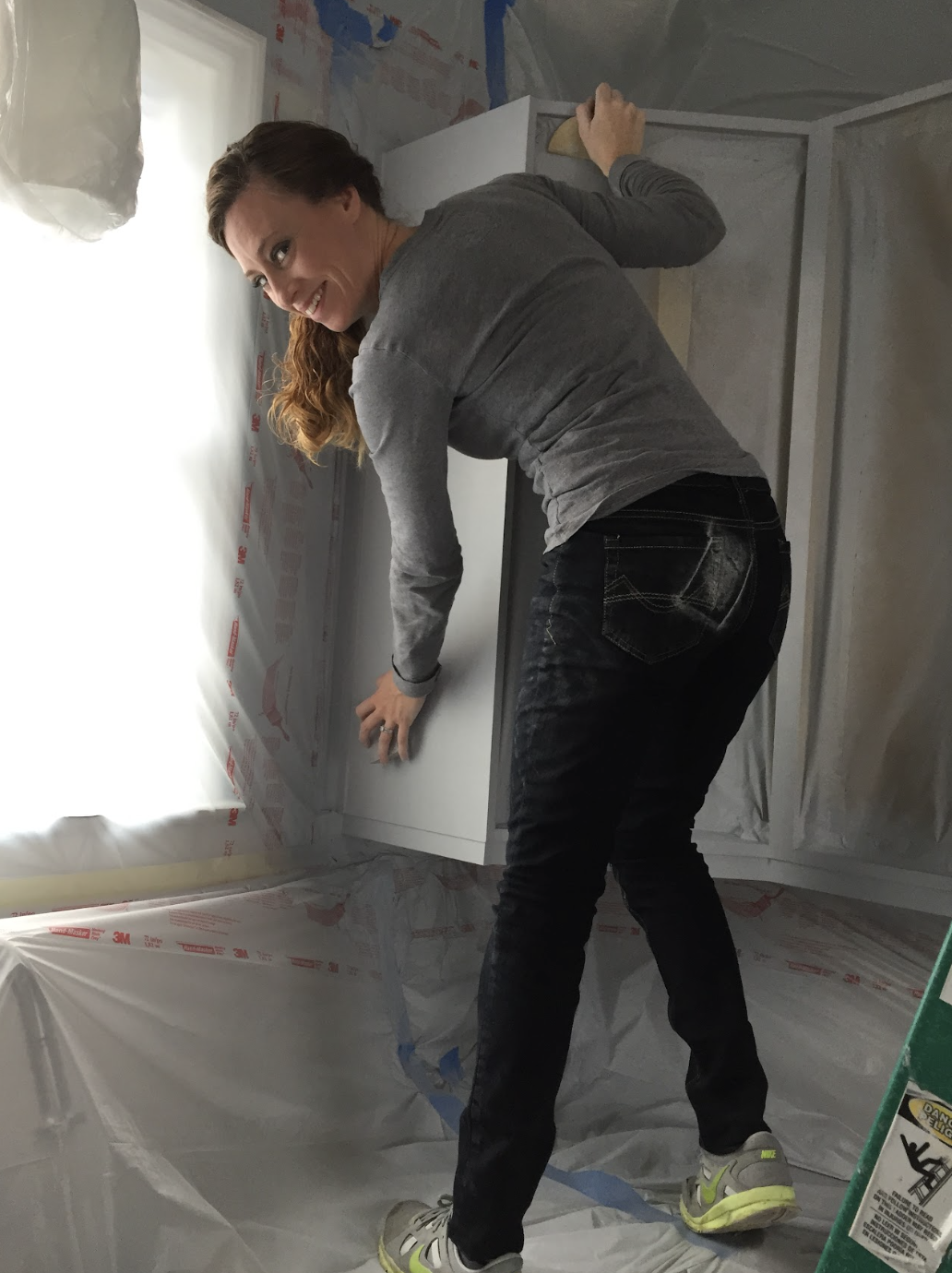 A woman is smiling while working on the installation of a cabinet in a room under renovation, surrounded by plastic sheeting and construction materials.