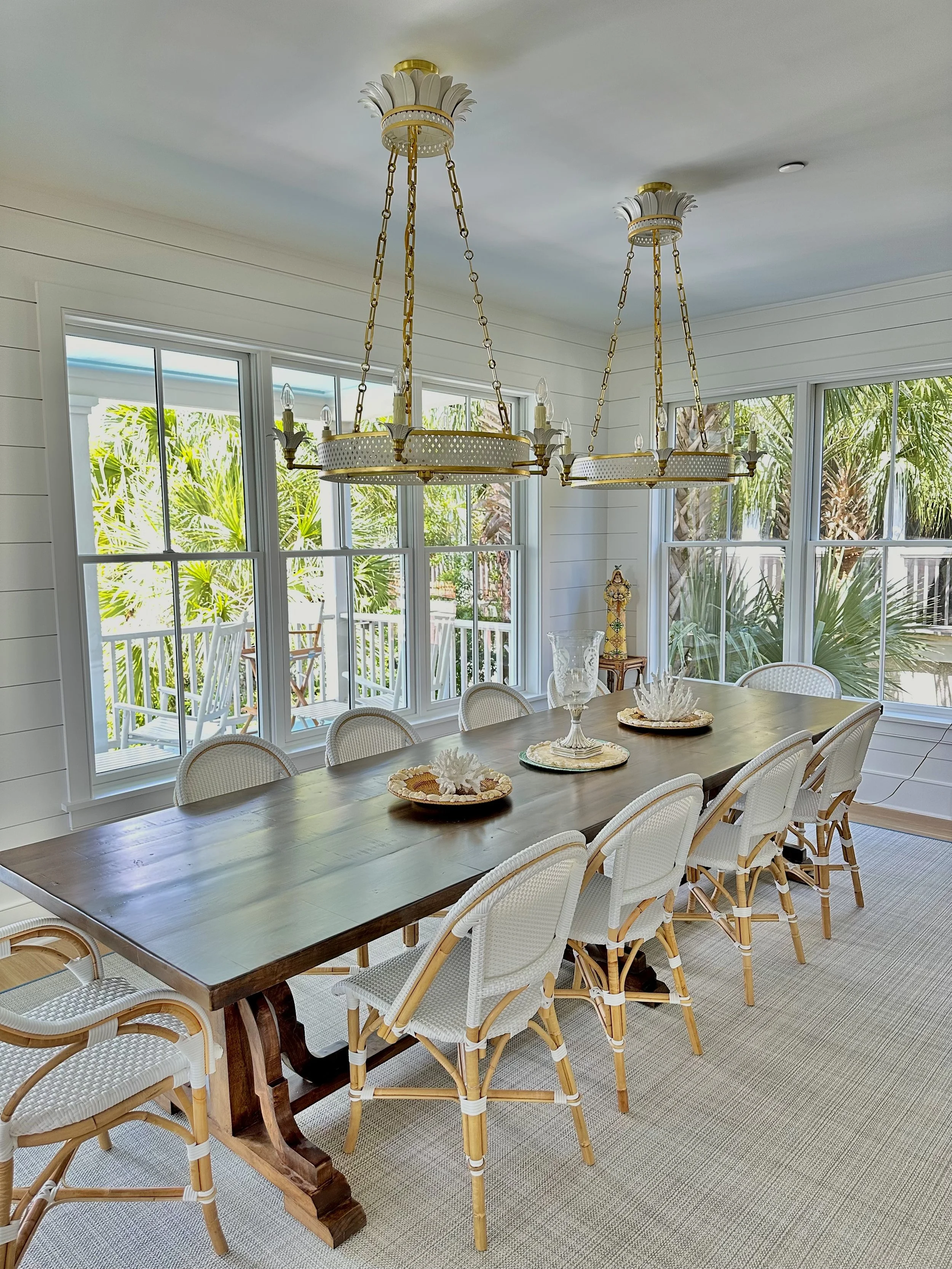 A dining room with a large wooden table surrounded by white wicker chairs, decorative trays and coral centerpiece, large windows showing greenery outside, and two gold chandeliers hanging from the ceiling.