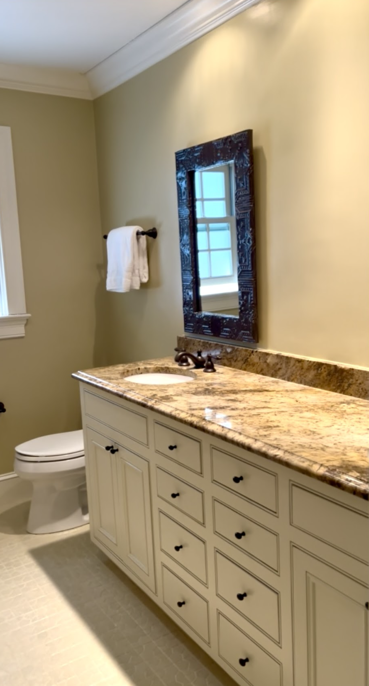 A bathroom featuring a white vanity with multiple drawers and black knobs, a granite countertop with a sink, a black-framed mirror, a window with a grid pattern, and a towel hanging on a black towel rod on a beige wall.
