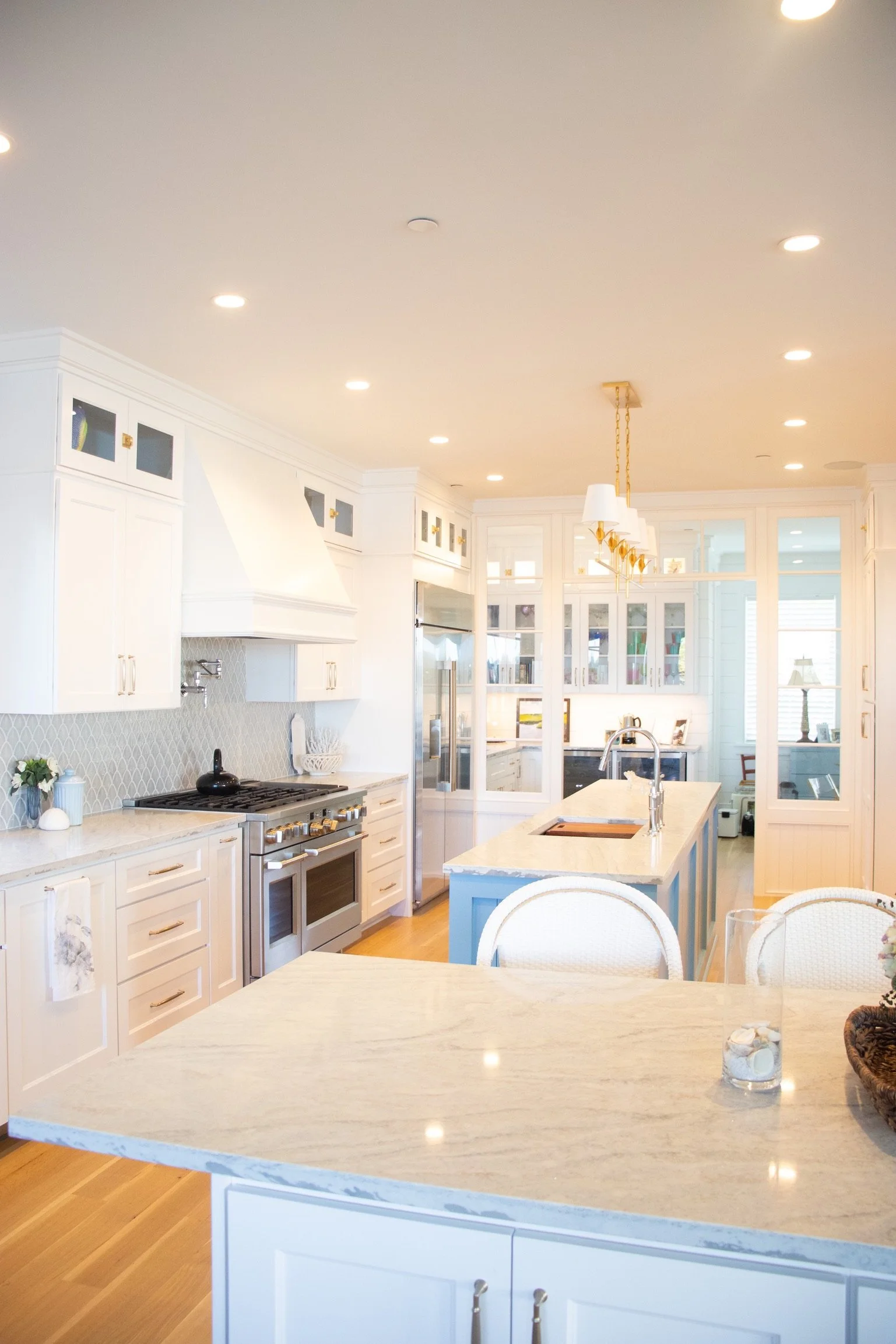 Bright, modern kitchen with white cabinetry, a marble island, and wooden floors. Recessed ceiling lights and a chandelier over the island. Glass cabinets, a stainless steel refrigerator, and a view into another room with a lamp and window.