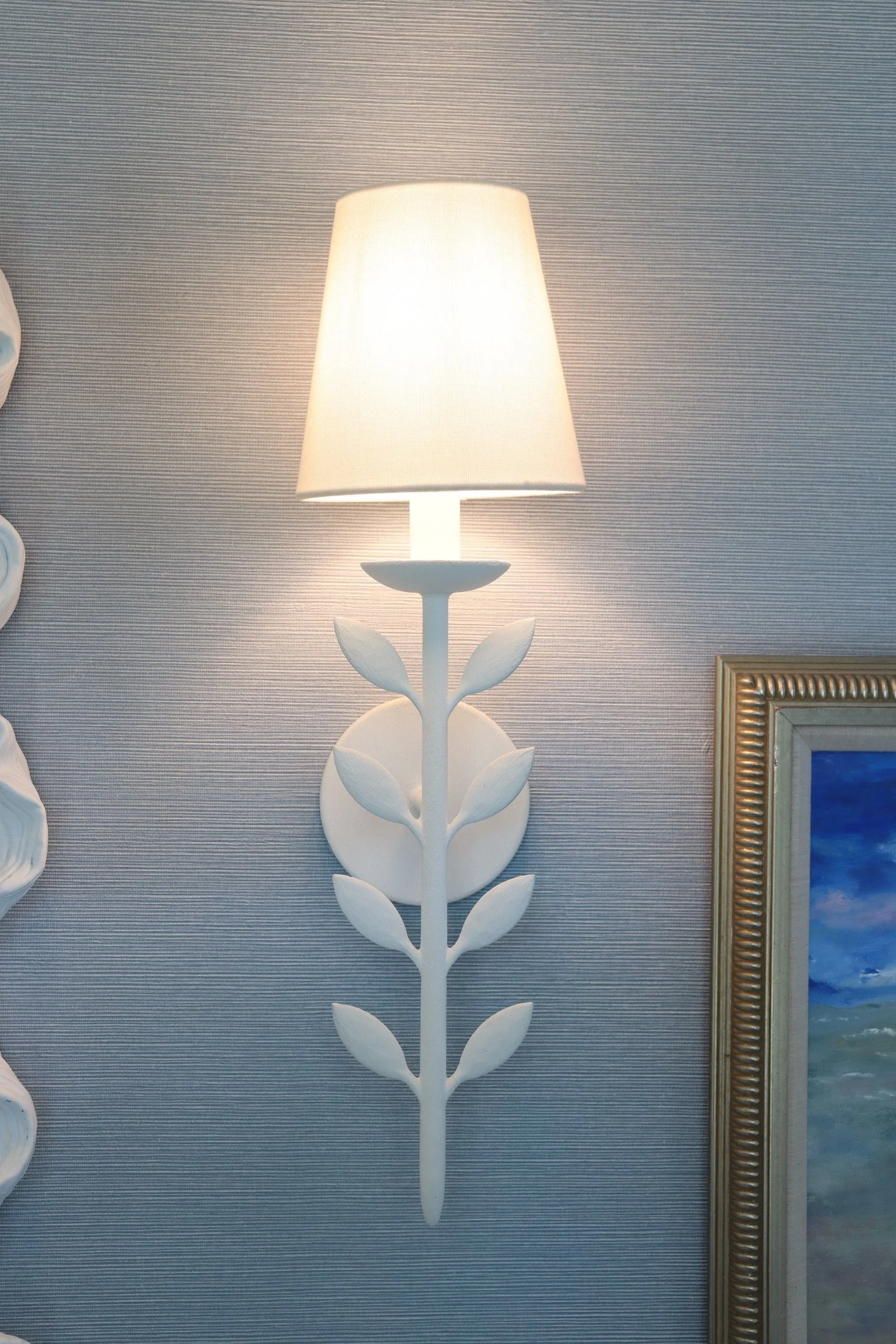 A white table lamp with a shade, mounted on a decorative floral-shaped stand, positioned against a textured wall with a framed painting partially visible on the right.