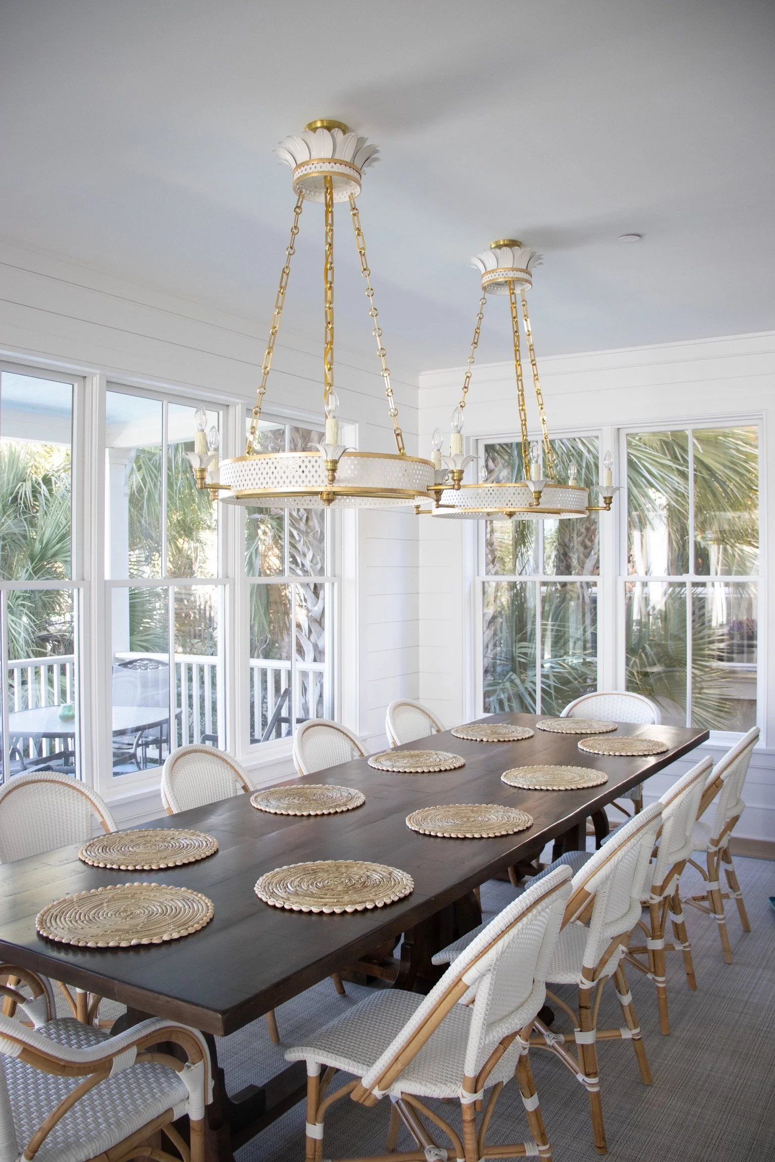 A dining room with a dark wooden table set with woven placemats, white wicker chairs, large windows showing palm trees outside, and two white and gold chandeliers hanging from the ceiling.