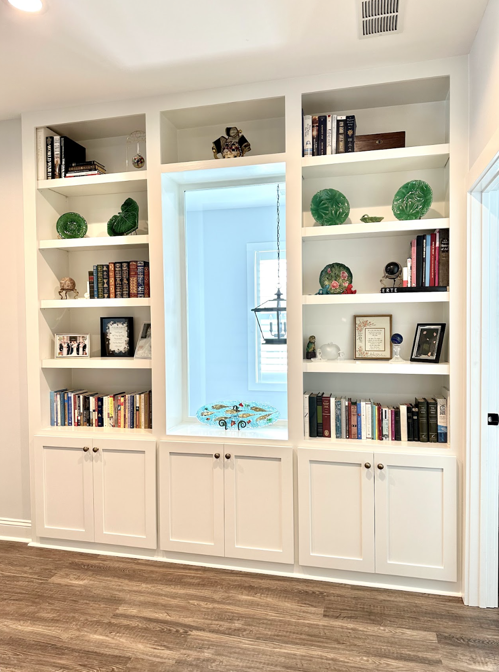 White built-in bookshelf with decorative green glass art and books, in a room with wooden flooring, small window with shutters, and a glass bowl on the ledge.