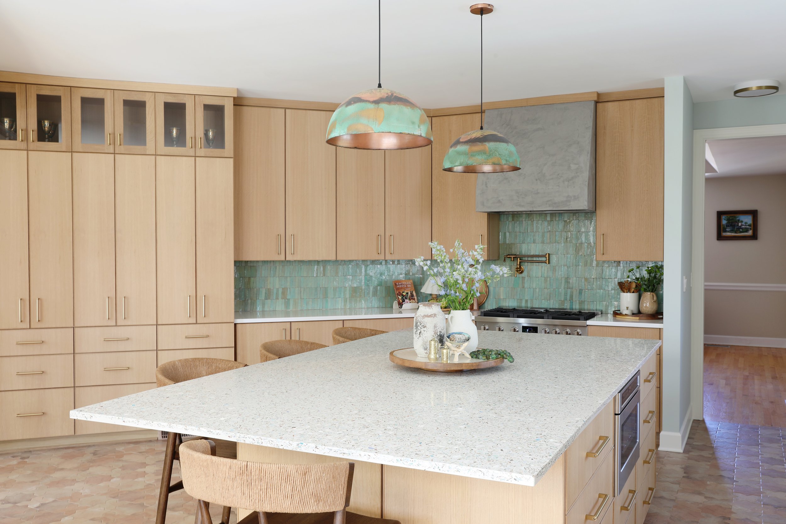 Modern kitchen with light wood cabinets, a large island with a speckled light countertop, and a green tile backsplash. Decorative items and flowers are on the island, with two artistic pendant lights hanging from the ceiling.